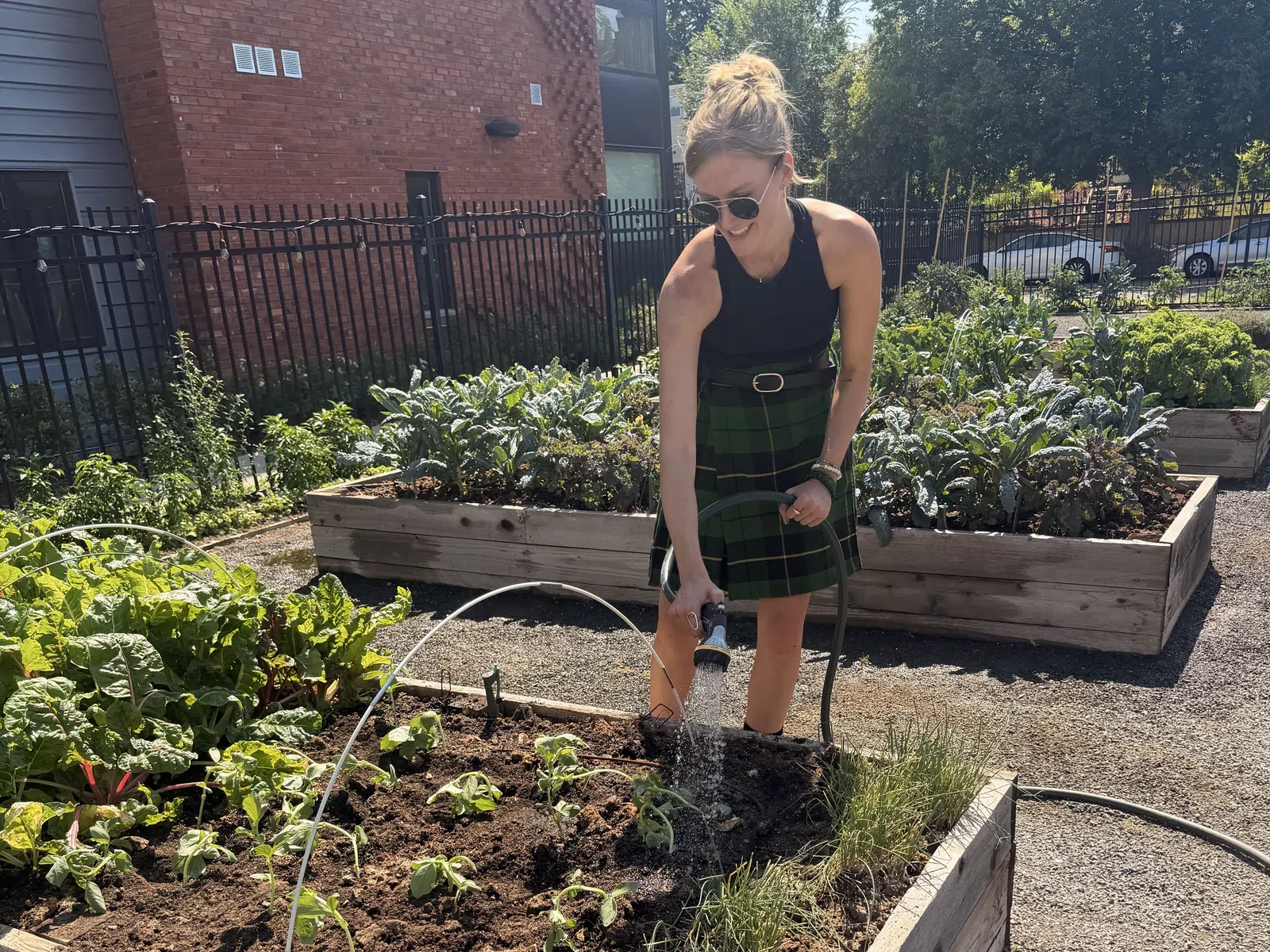 Woman watering plants in a raised wooden garden bed outside on a sunny day near brick building.