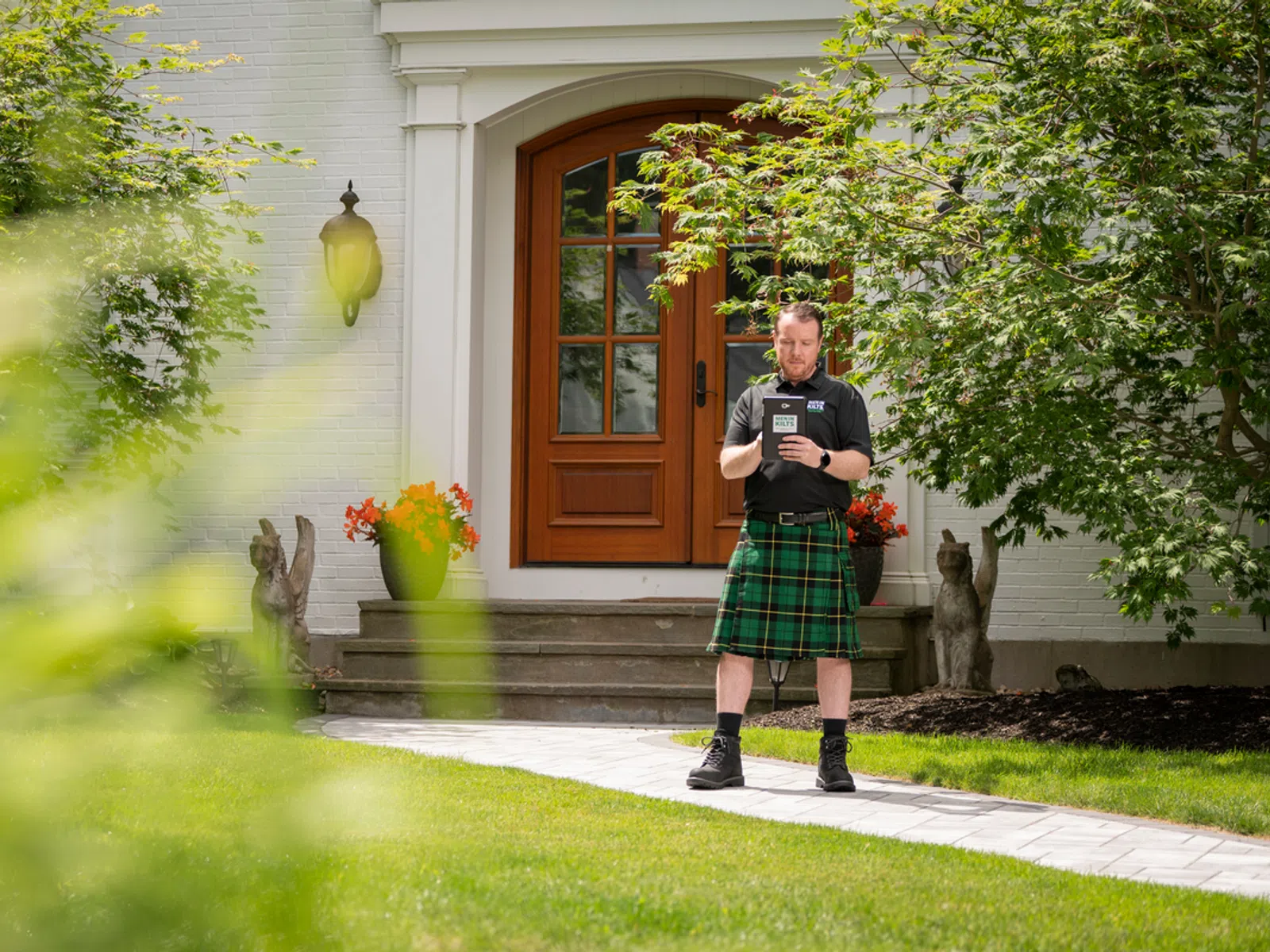 Man in green plaid kilt stands on walkway in front of house with wooden door and garden statues.