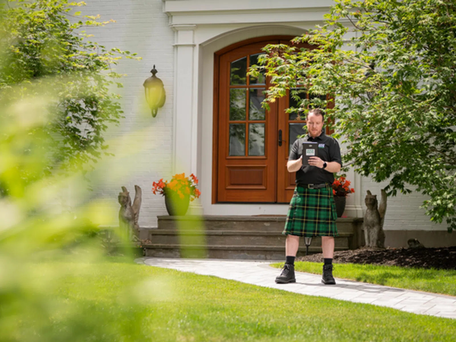 Man in green plaid kilt stands on walkway in front of house with wooden door and garden statues.