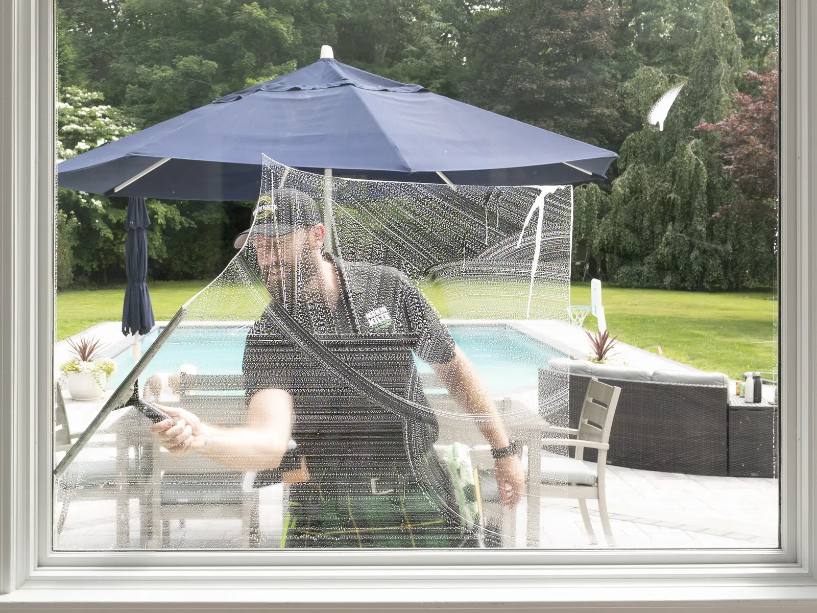 Window cleaner using a squeegee to wash large glass window by outdoor patio with umbrella and pool