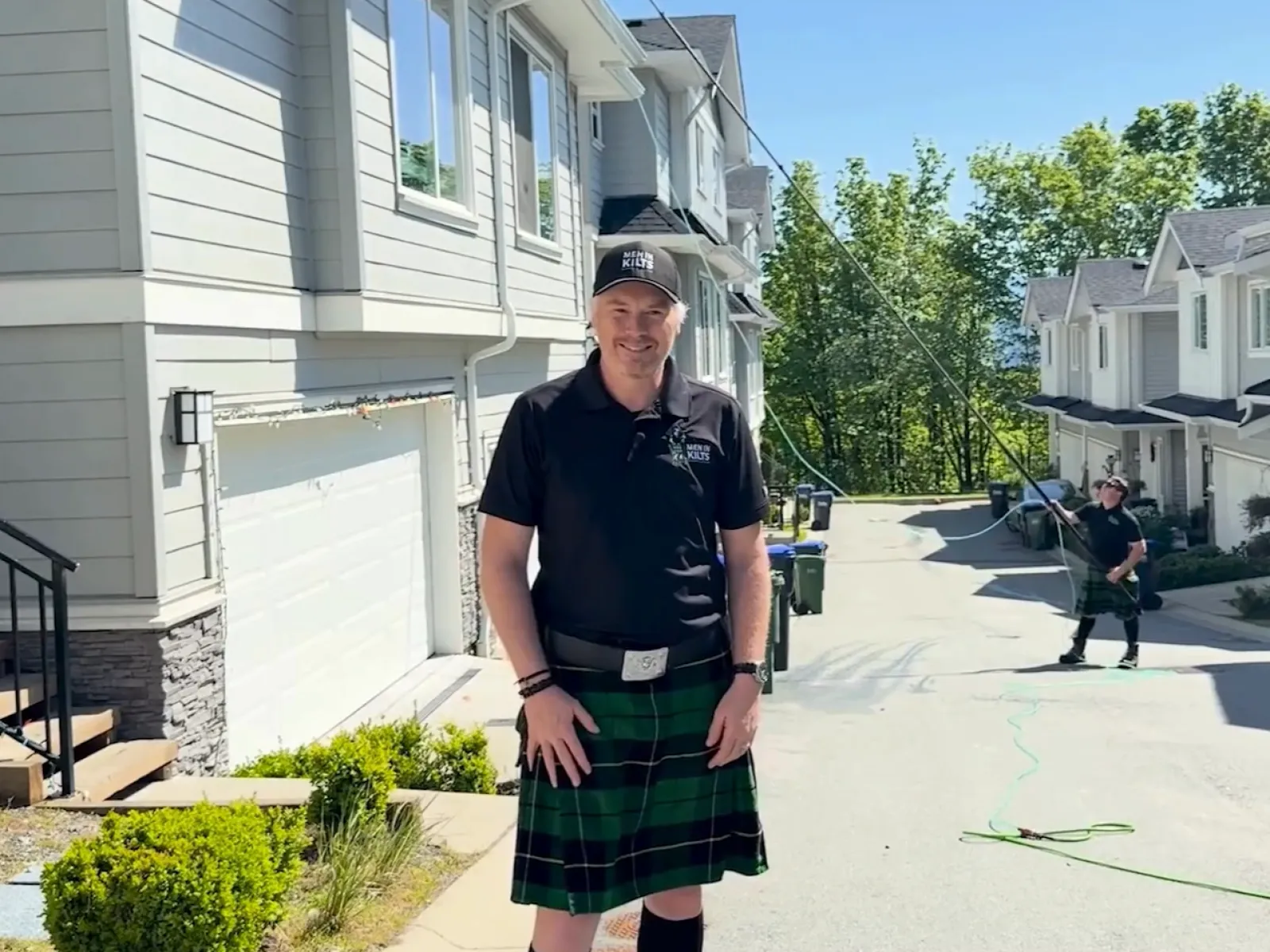 Man in a black shirt and green kilt standing on a sunny suburban street with houses and trees in the background.