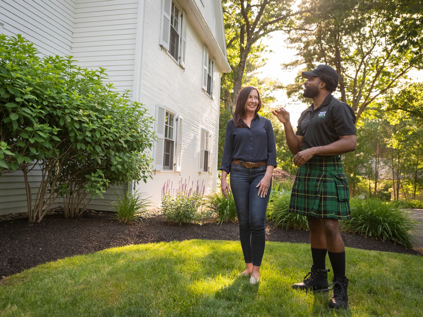 A homeowner speaks with a landscaper in a green lawn, surrounded by vibrant bushes near a white house.
