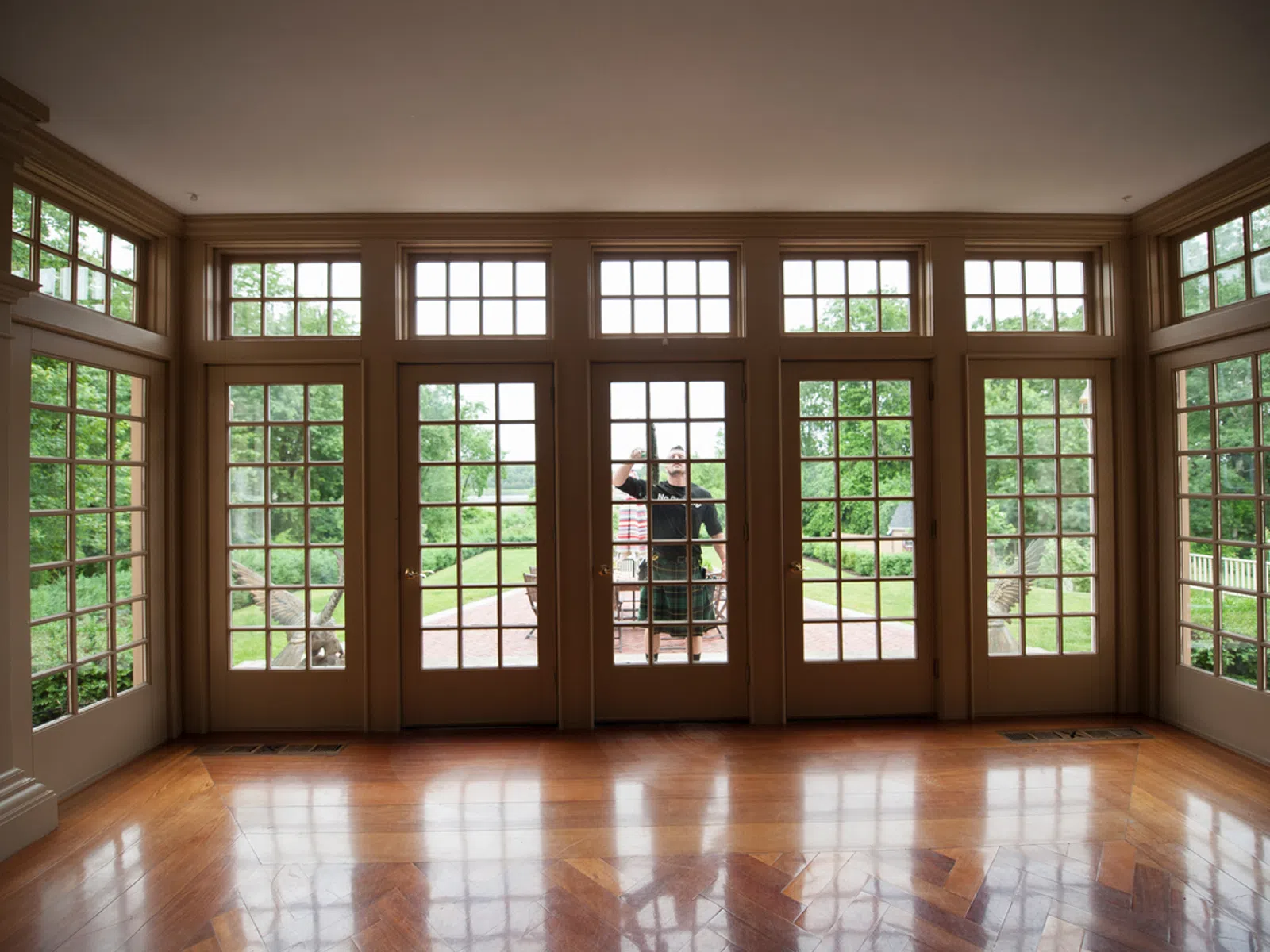 Man in kilt cleaning large glass doors in a spacious room with wooden floor and garden view outside.