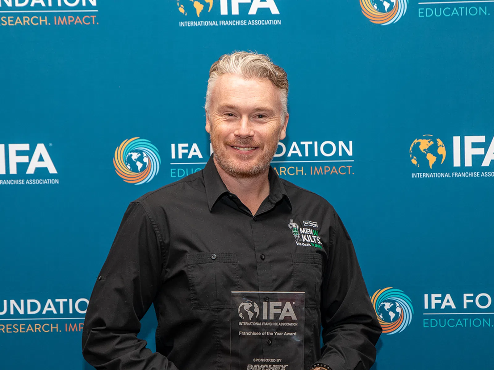 Man wearing a kilt and black shirt holding IFA Franchisee of the Year Award in front of IFA Foundation backdrop
