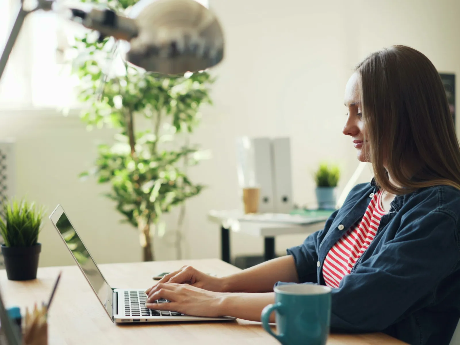 Young woman typing on laptop at bright desk with green plant and blue mug in modern home office