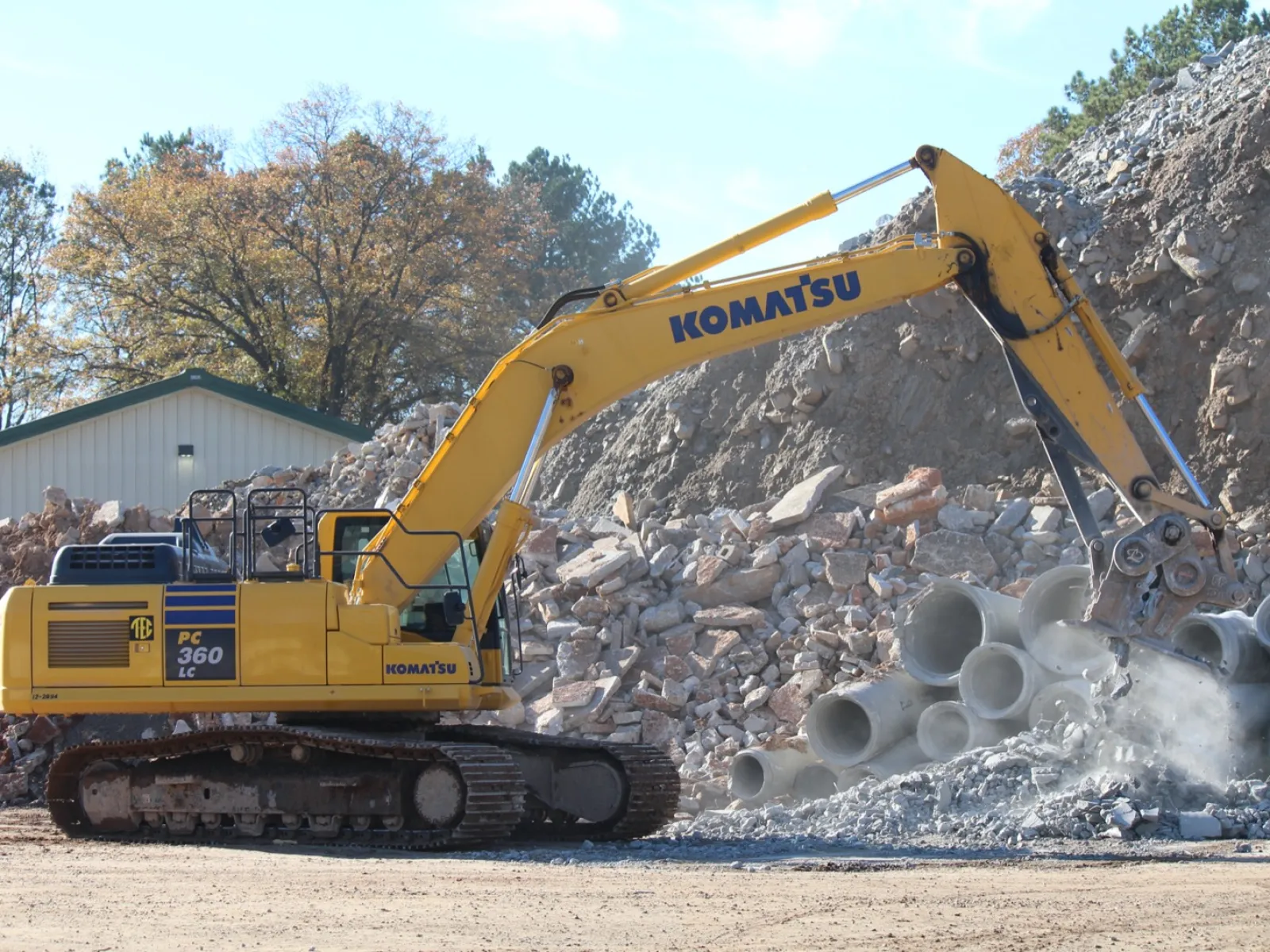 Yellow Komatsu PC360 excavator demolishing concrete pipes at a construction site with rubble and trees nearby