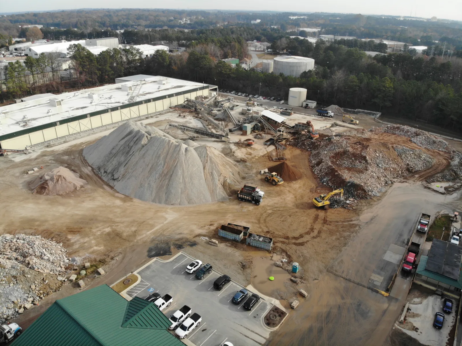 Aerial view of industrial construction site with dirt piles, heavy machinery, trucks, and parking area near warehouse buildings.