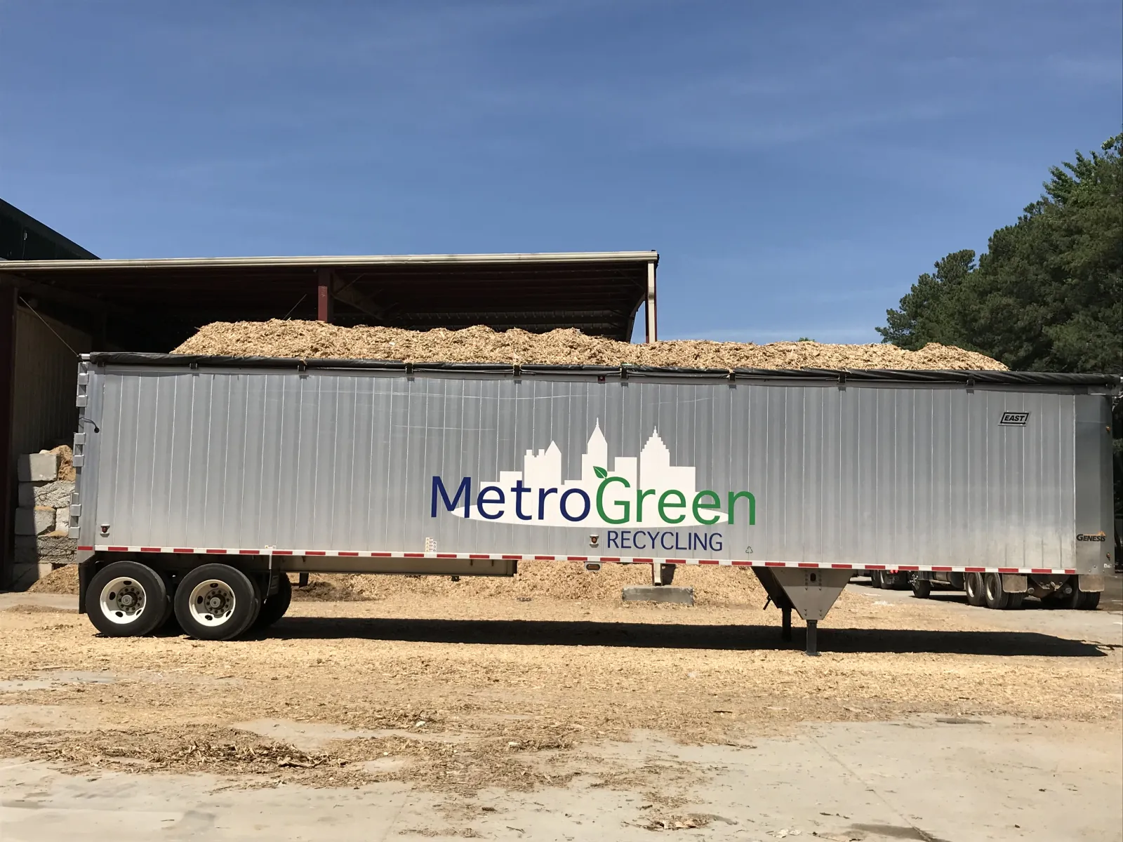 Metal recycling trailer with MetroGreen Recycling logo loaded with wood chips under a clear blue sky.