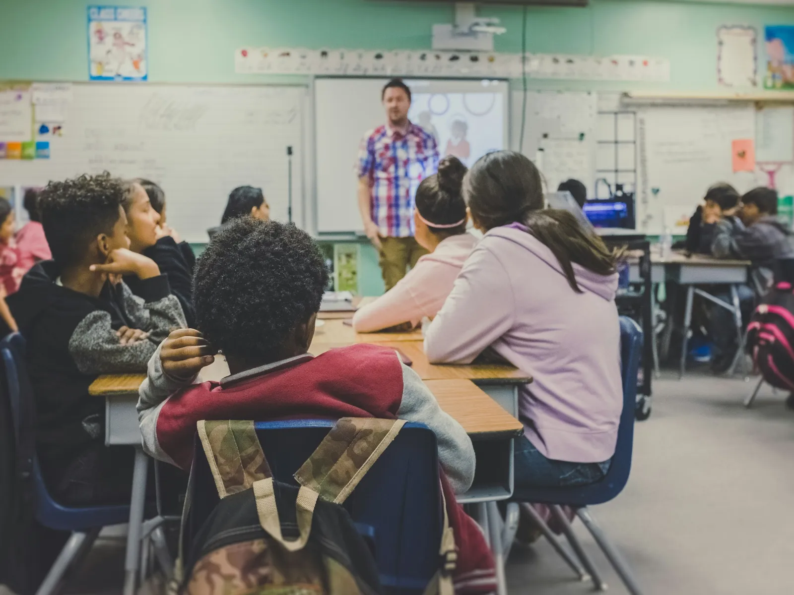 Students in a classroom attentively listening to a teacher giving a lesson at the front of the room.