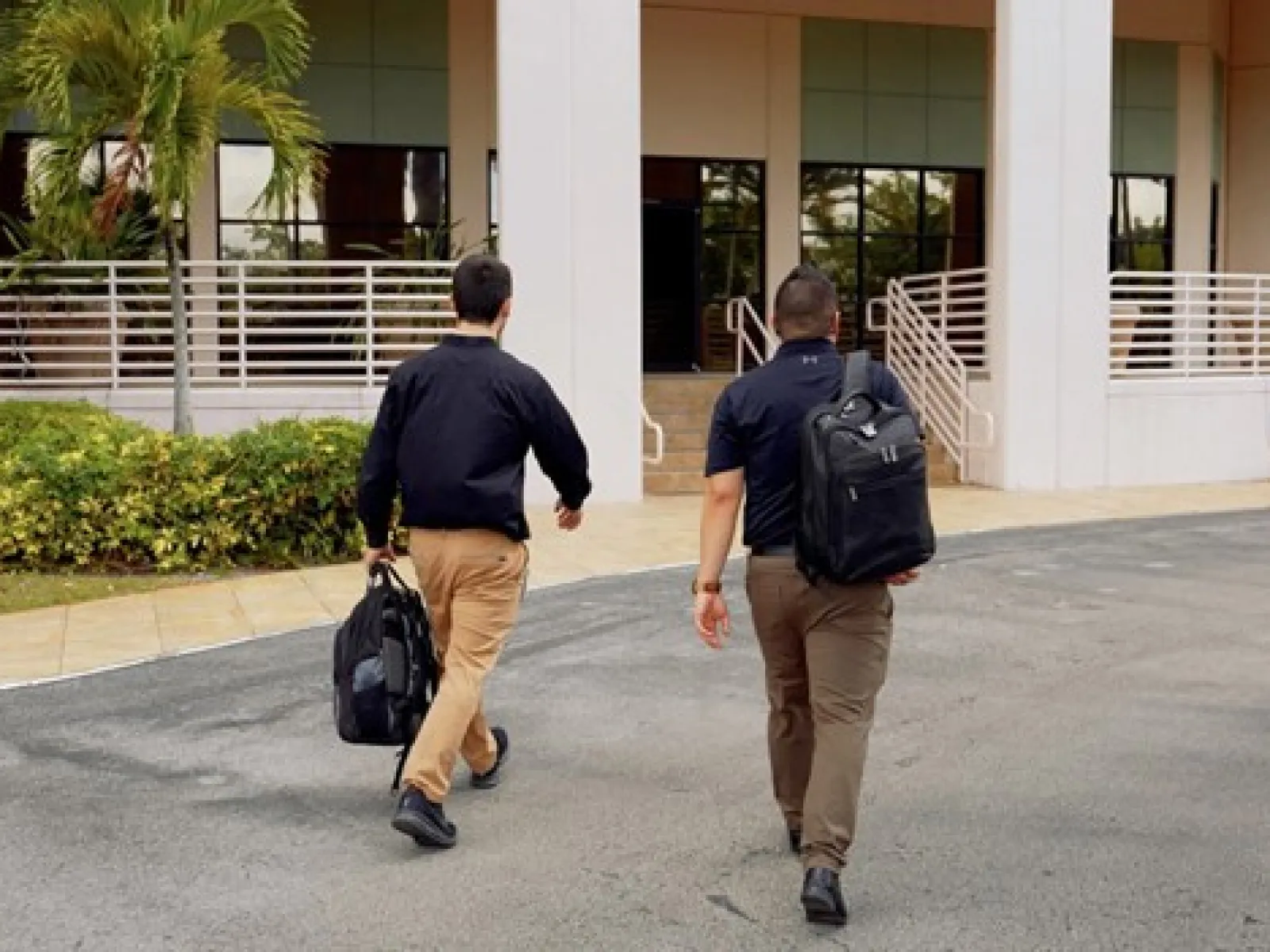 Two men with backpacks walking toward a modern building entrance surrounded by greenery and palm trees.
