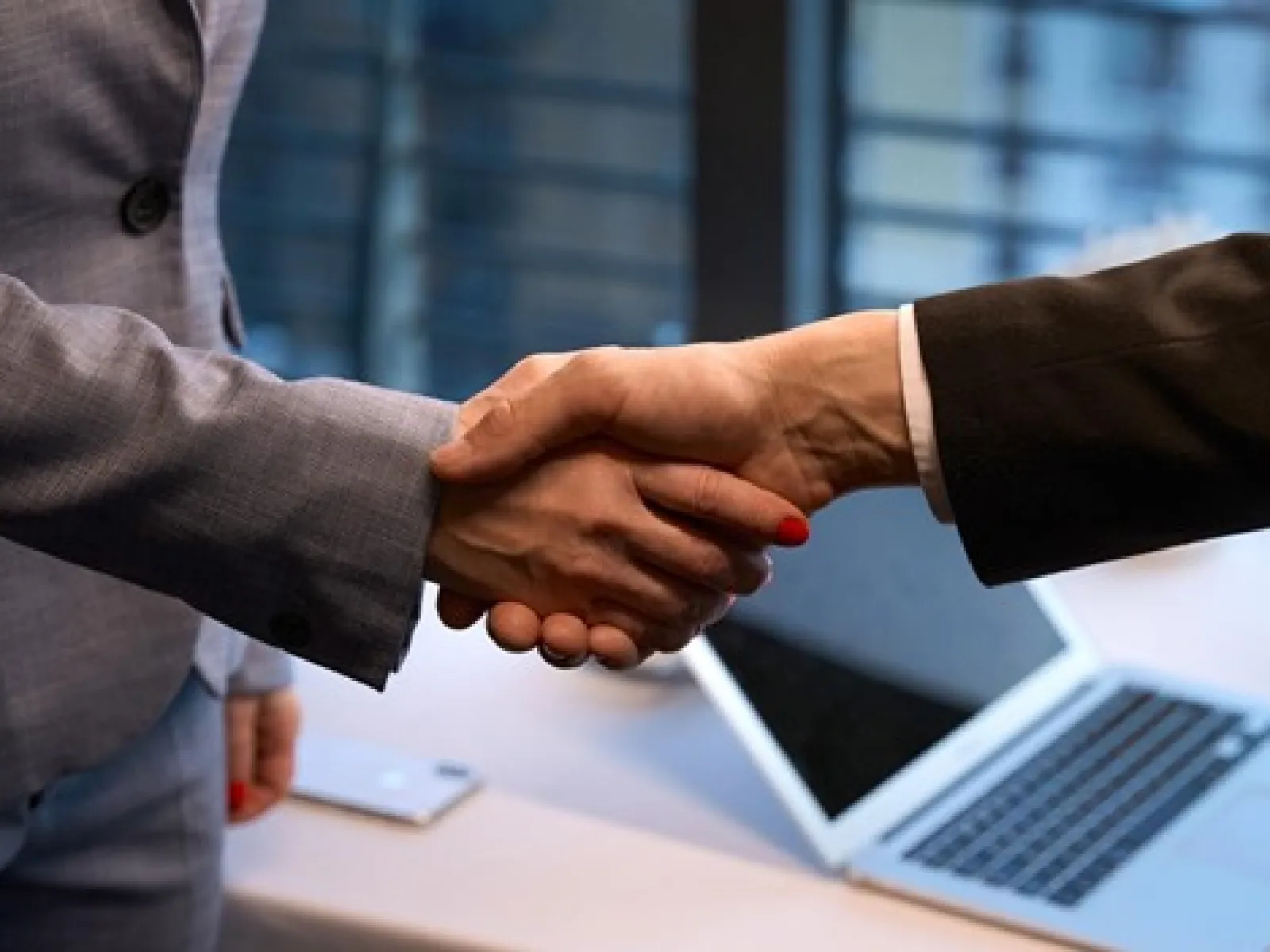 Two business professionals shaking hands over a desk with a laptop in a modern office setting.