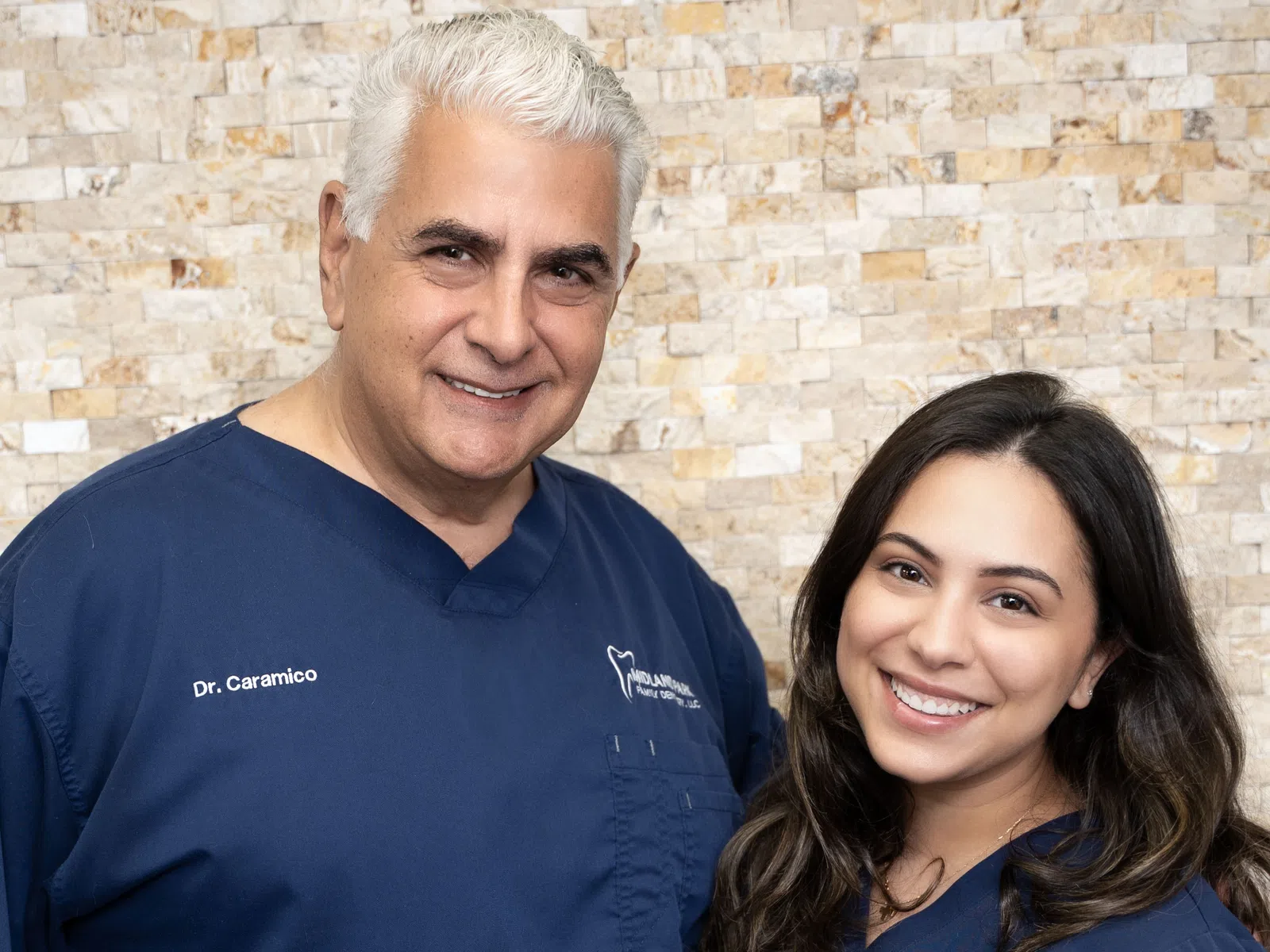 Smiling male and female dentists in blue scrubs standing against a beige stone wall background.