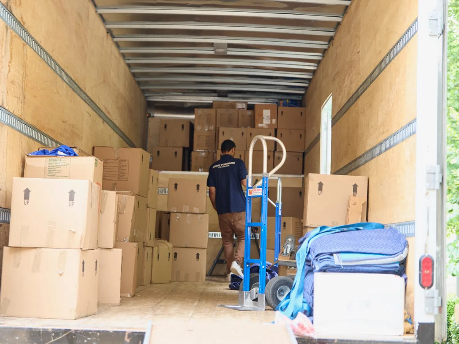 Worker unloading packed cardboard boxes inside a moving truck with a hand truck and moving blankets.