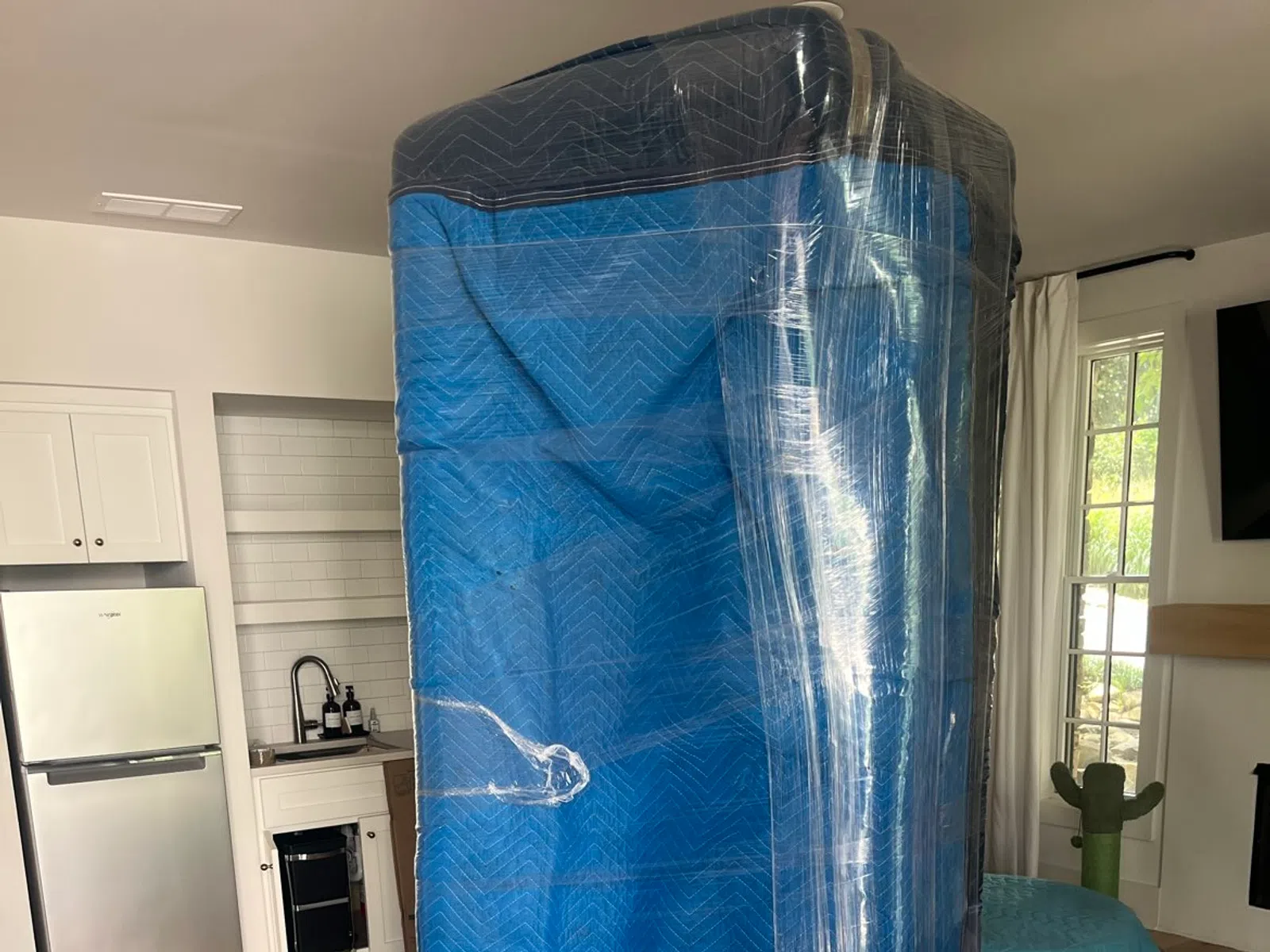 Large blue cushioned furniture wrapped in protective plastic film standing on wooden floor indoors.