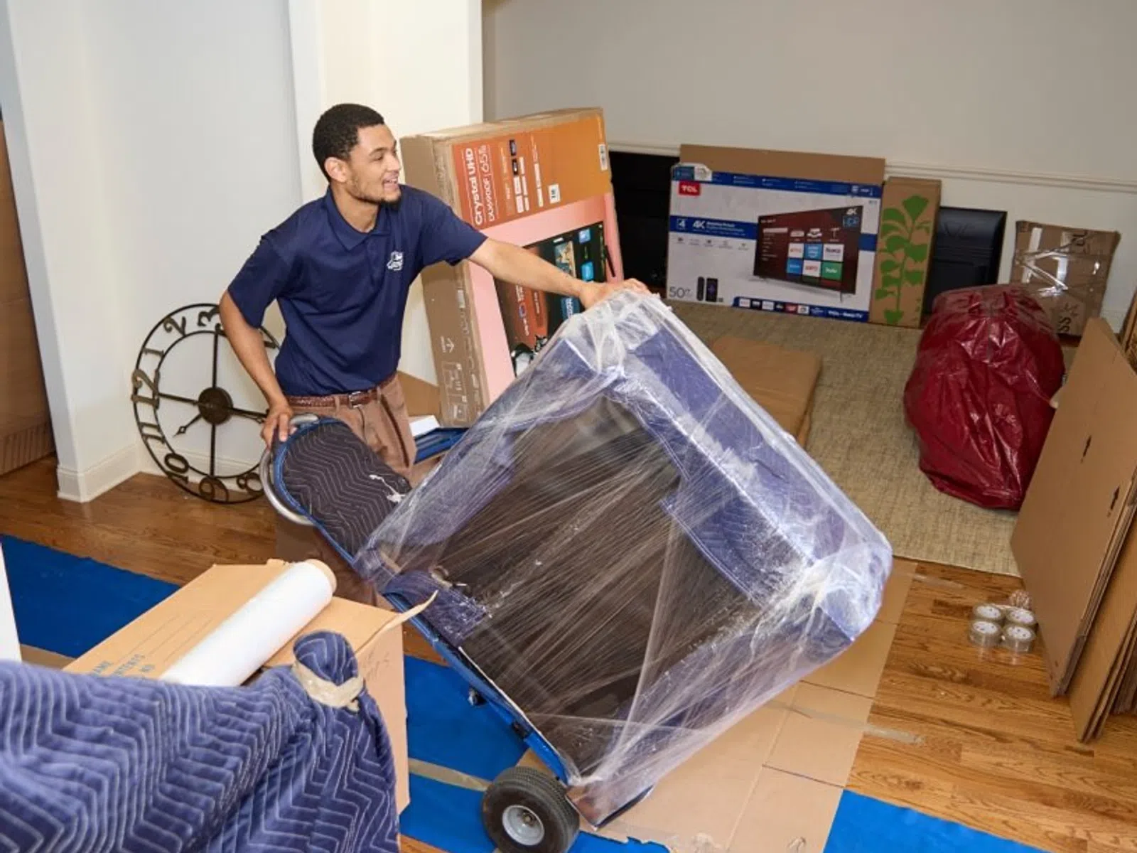 Mover transporting wrapped furniture on a dolly inside a home surrounded by packed boxes and protective coverings.
