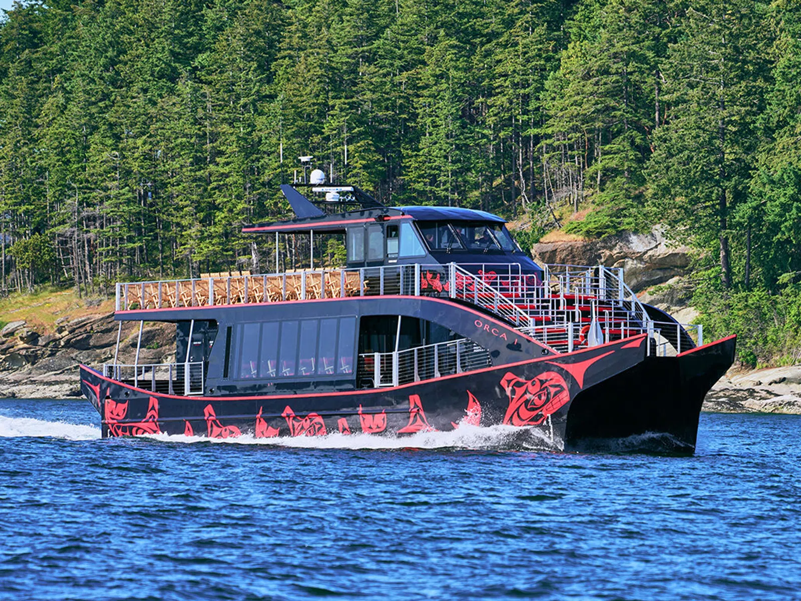 A vibrant black and red tour boat with native art cruising through a scenic waterway surrounded by lush greenery.