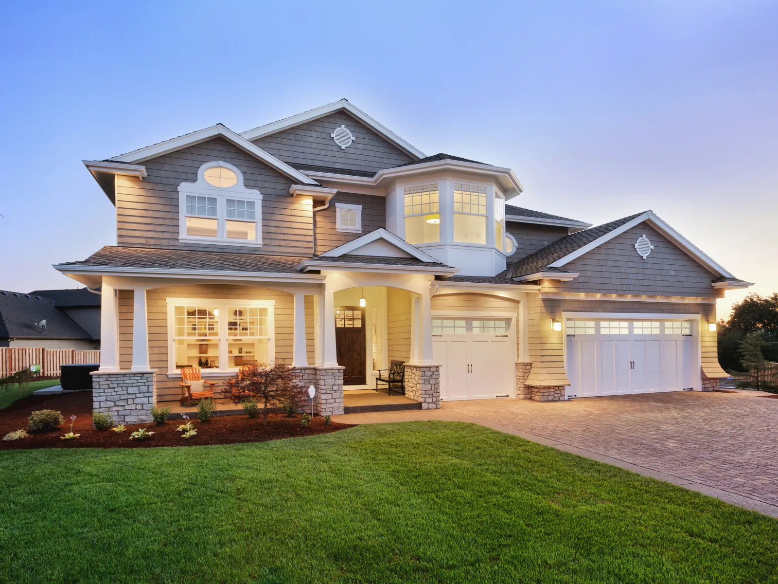 Modern two-story house with large garage, stone pillars, porch, and manicured lawn at dusk with exterior lights on