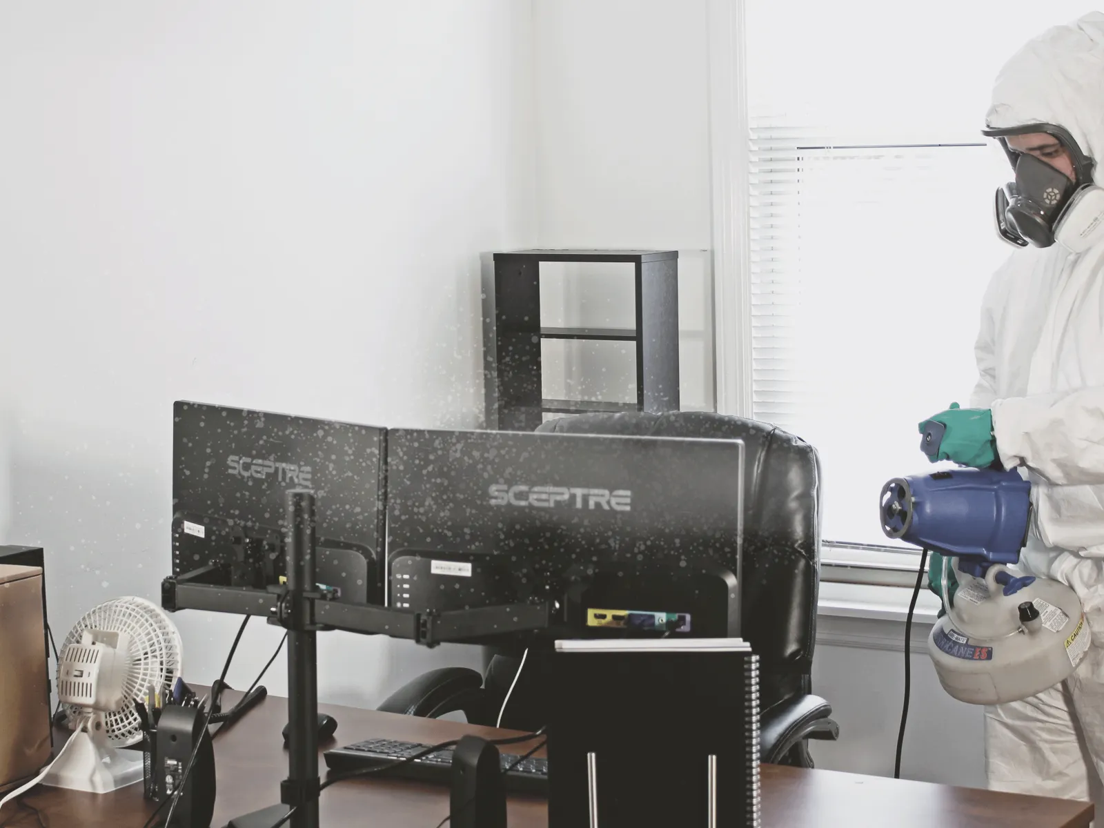 Person in protective suit and mask disinfecting office workspace with computers and a fan near window.