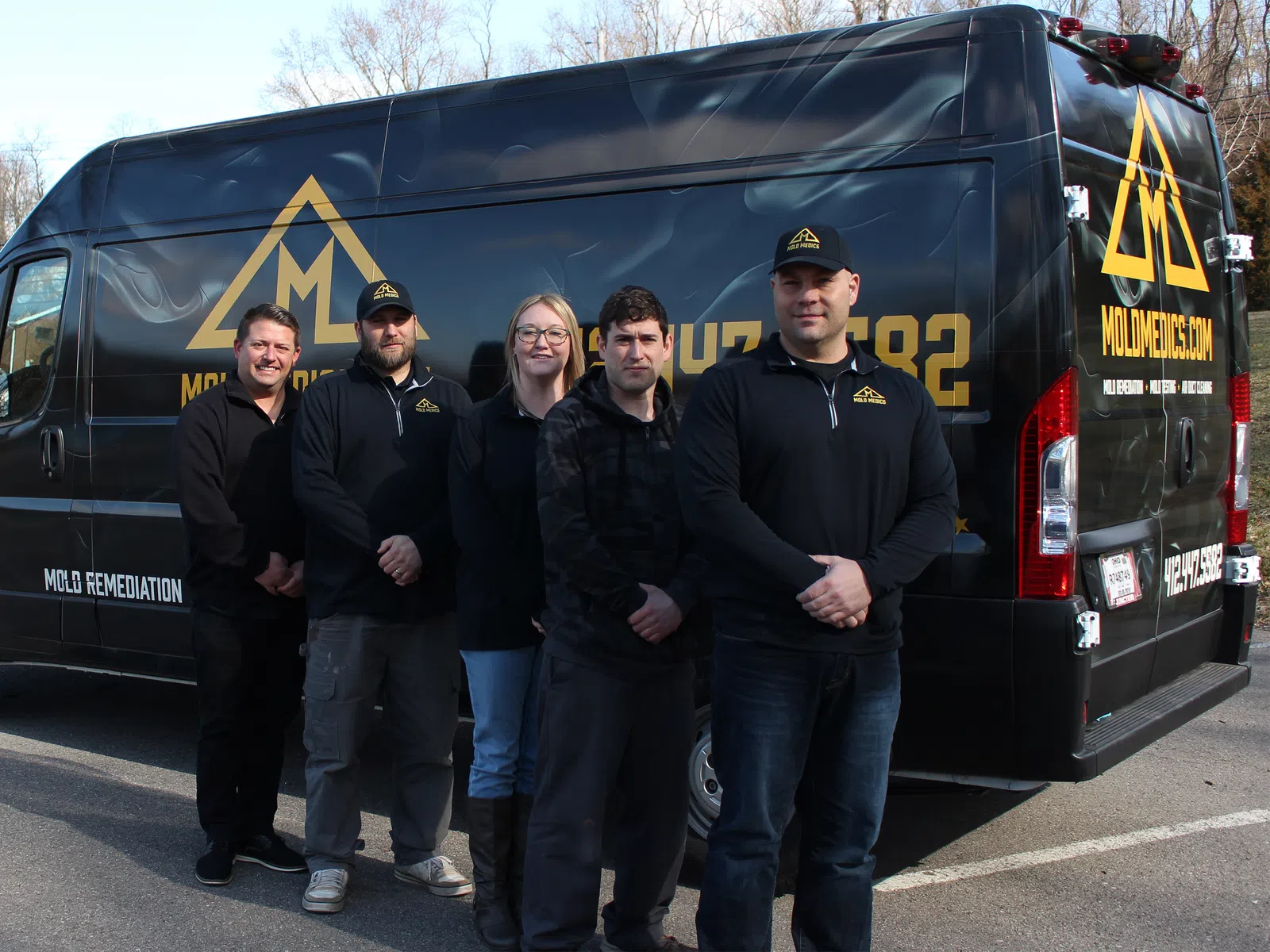 Group of five mold remediation professionals standing in front of a branded black company van with yellow logo.