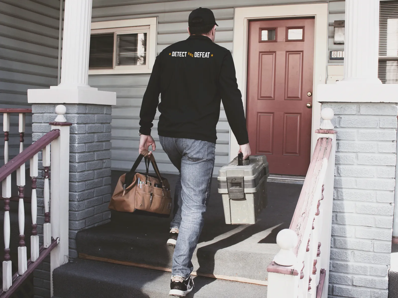 Technician carrying toolboxes walking up porch steps toward a front door of a residential home.