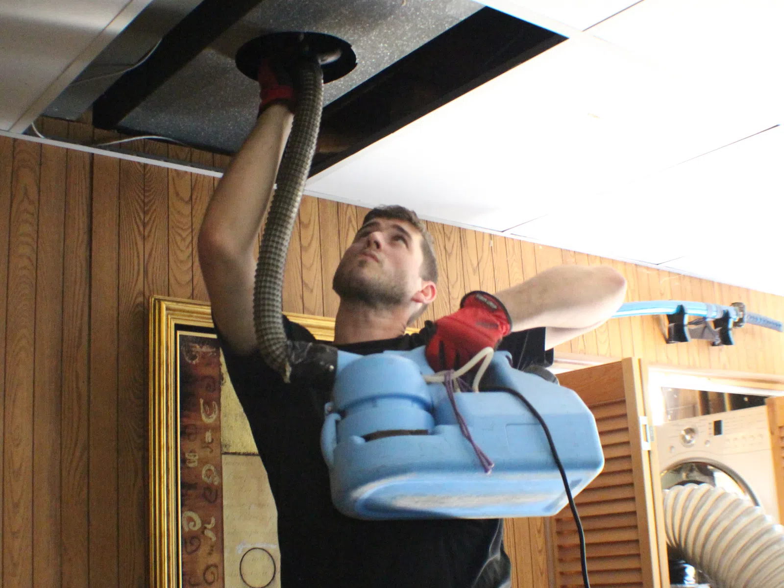 Technician wearing gloves performs duct cleaning using vacuum equipment in wooden paneled room.