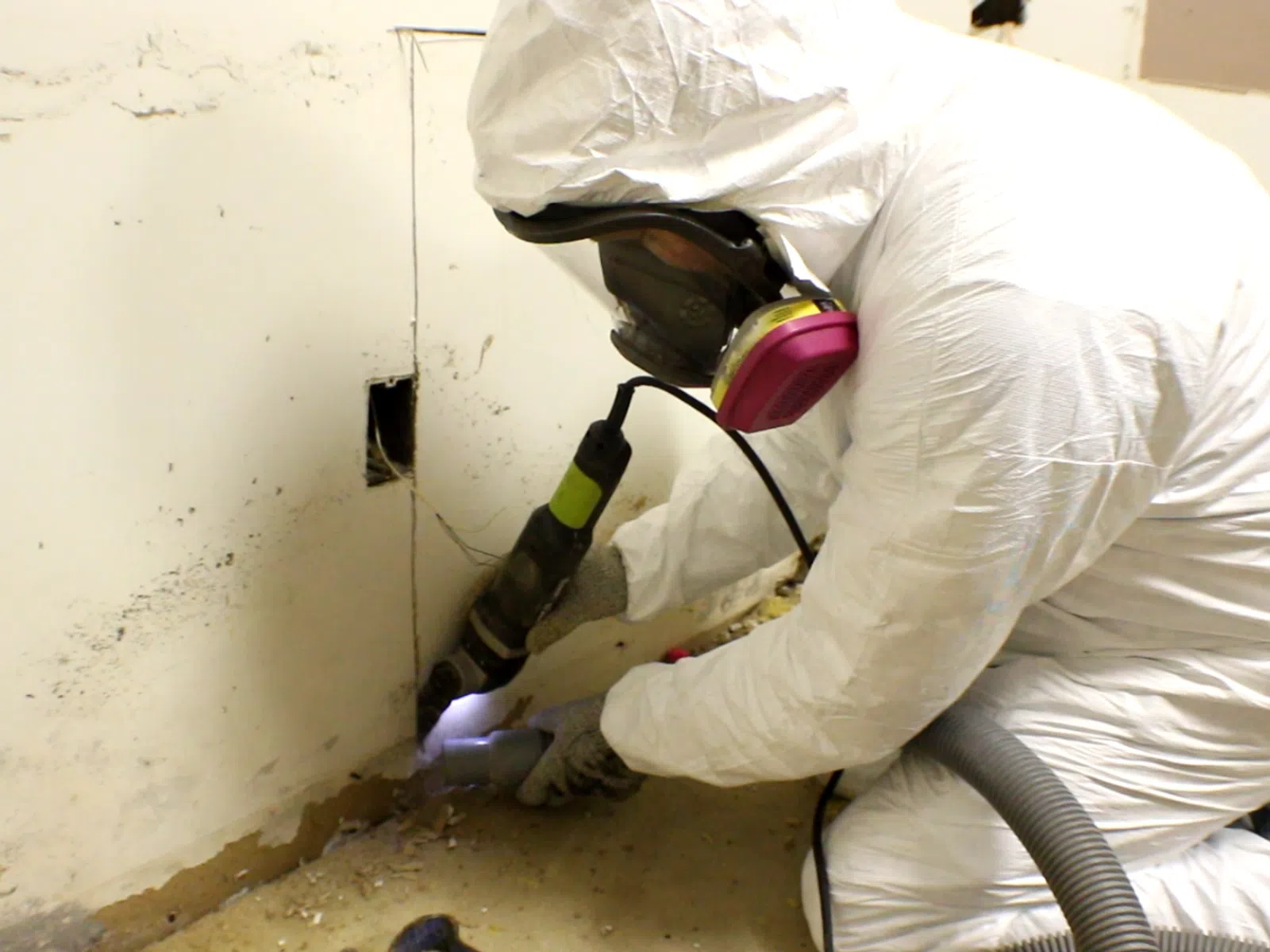 Technician in protective suit and respirator removing mold from a damaged wall corner using power tools indoors.