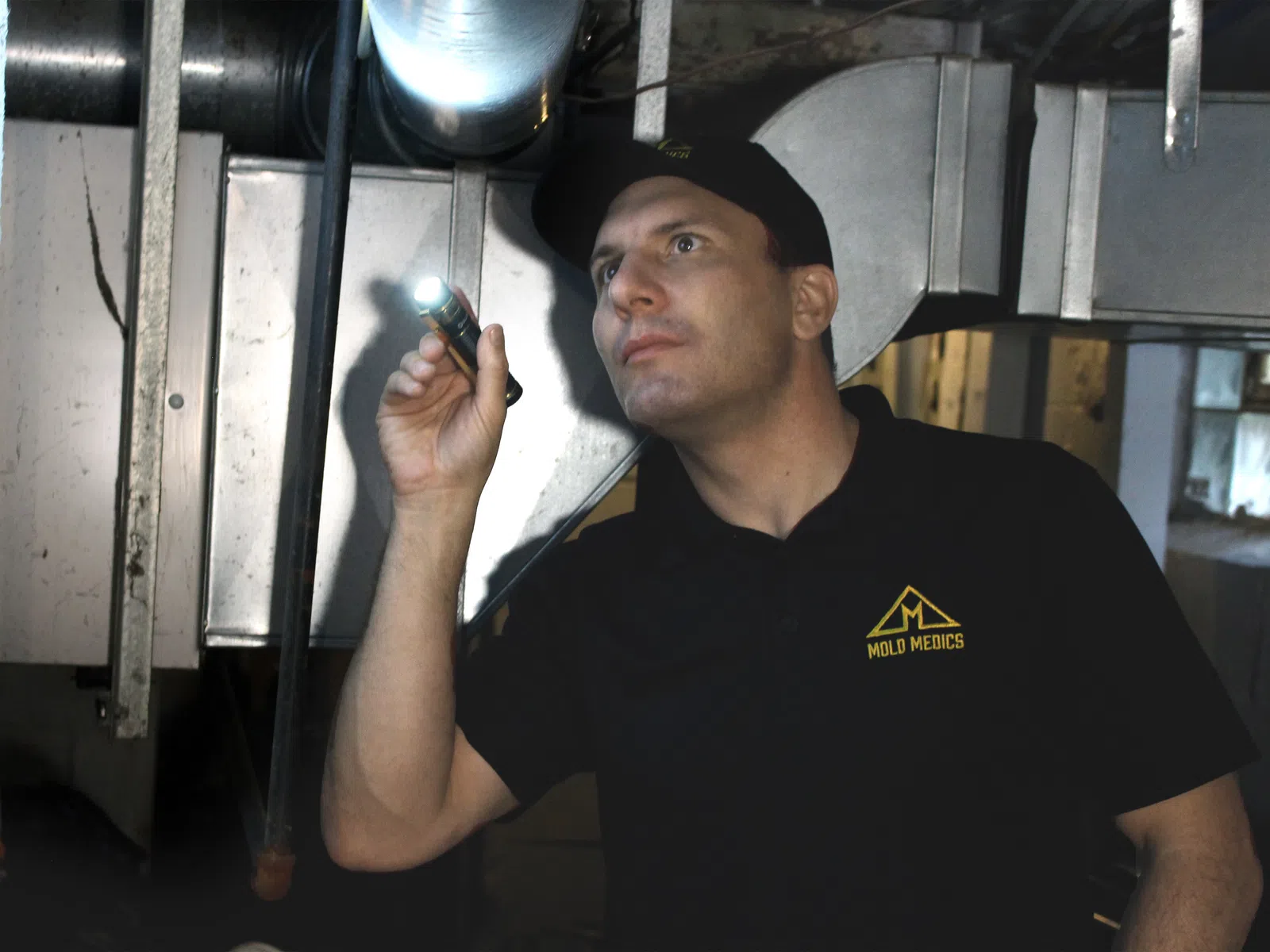 Technician in Mold Medics uniform inspecting HVAC ductwork in a basement with a flashlight.