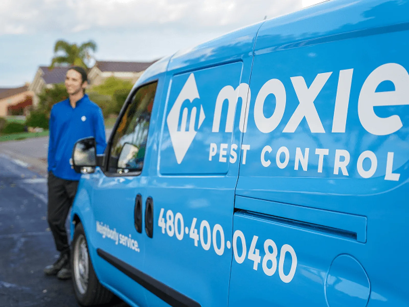 Blue Moxie Pest Control van parked on street with technician in uniform standing nearby under cloudy sky
