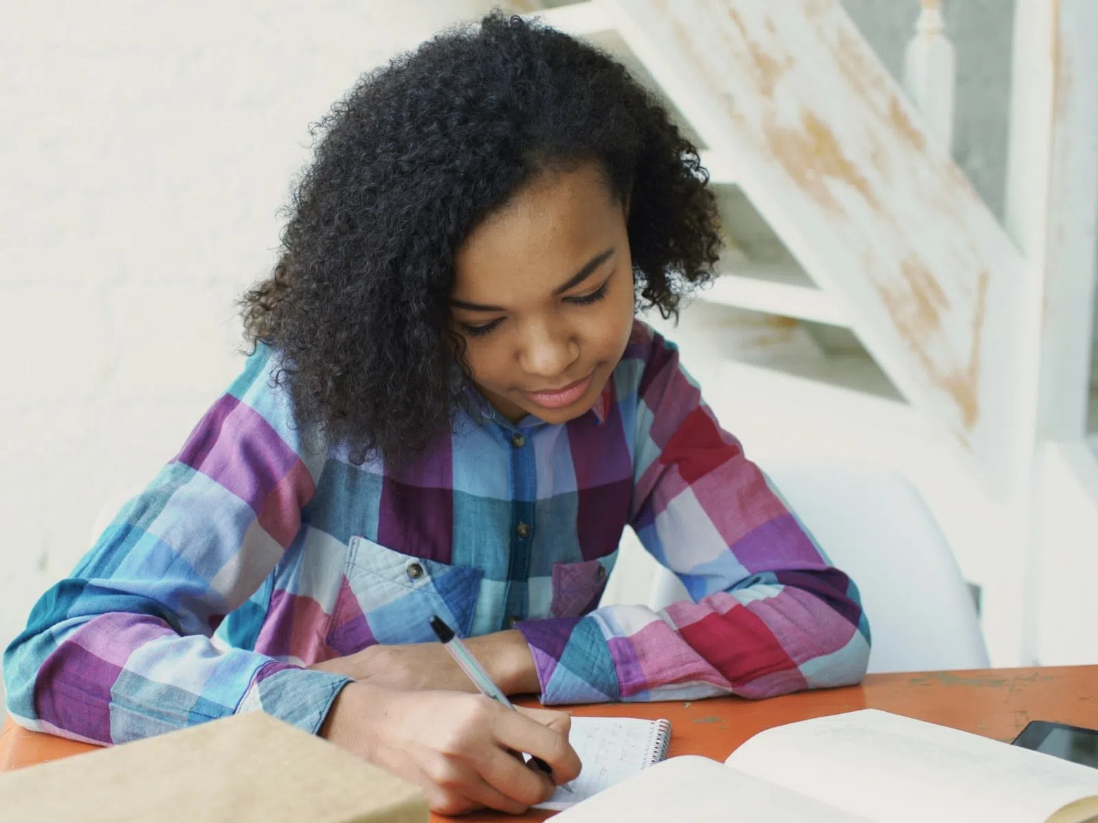 Young girl in a colorful plaid shirt writing in a notebook at a wooden table with books and a tablet