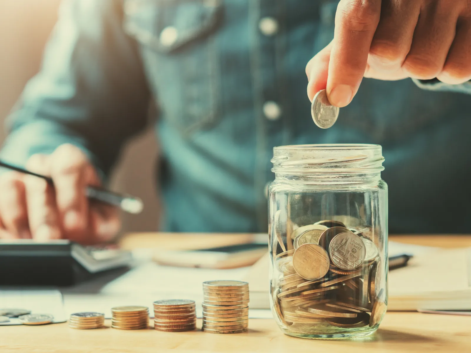Person placing coin in jar with stacked coins and calculator on table, representing saving money.