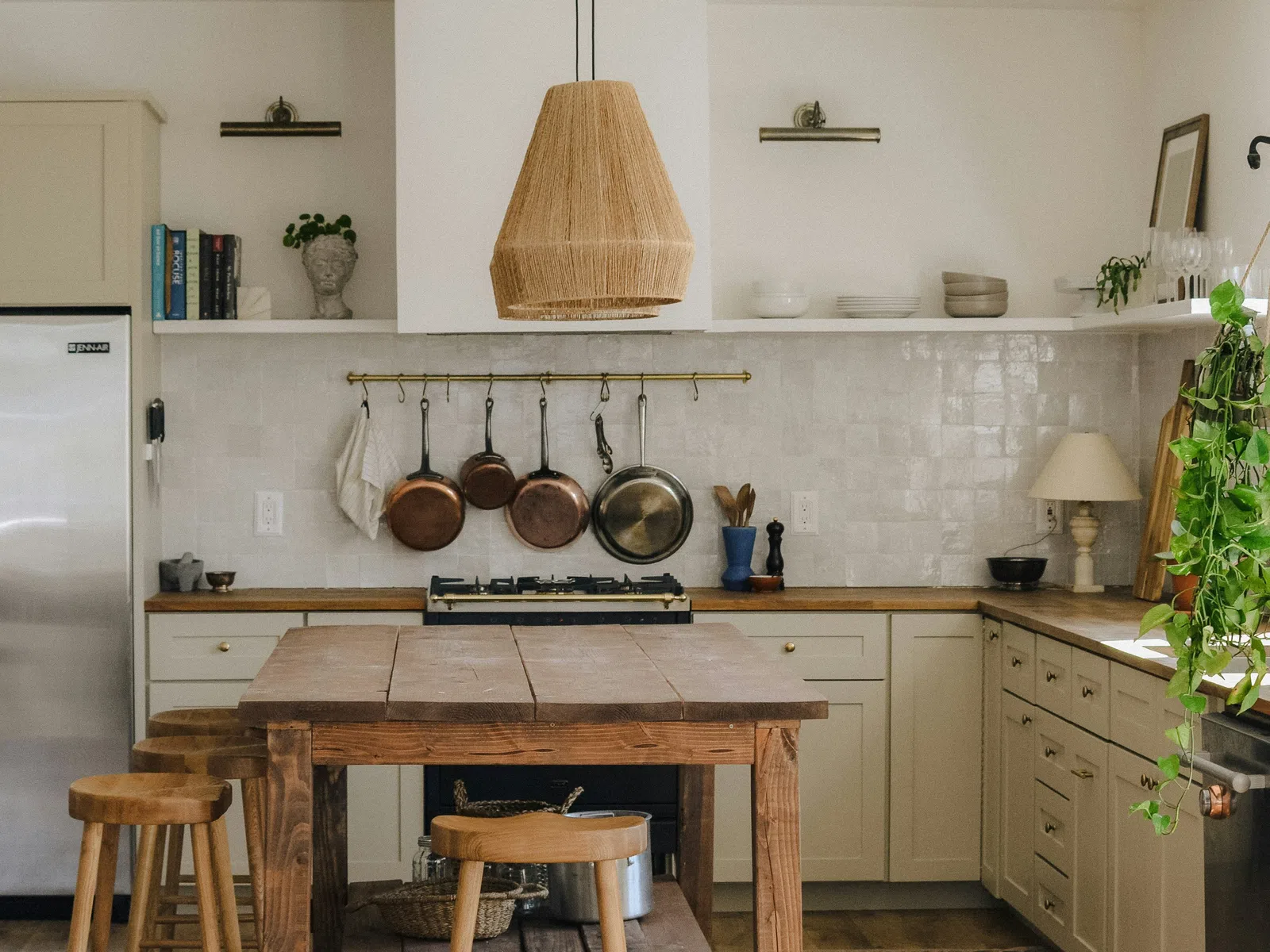Bright farmhouse kitchen with wooden island, stools, hanging copper pans, and wicker pendant light fixture.