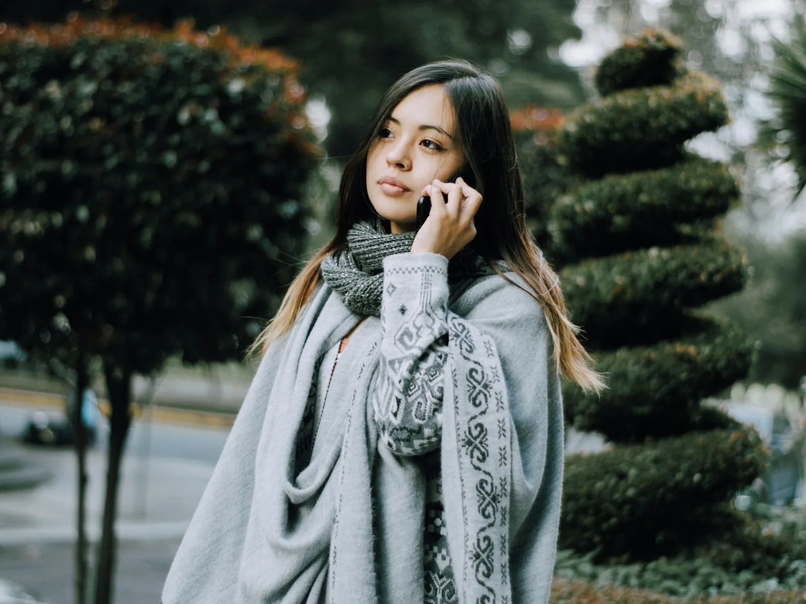 Young woman wearing a gray patterned sweater and scarf talking on a smartphone outdoors in a park.