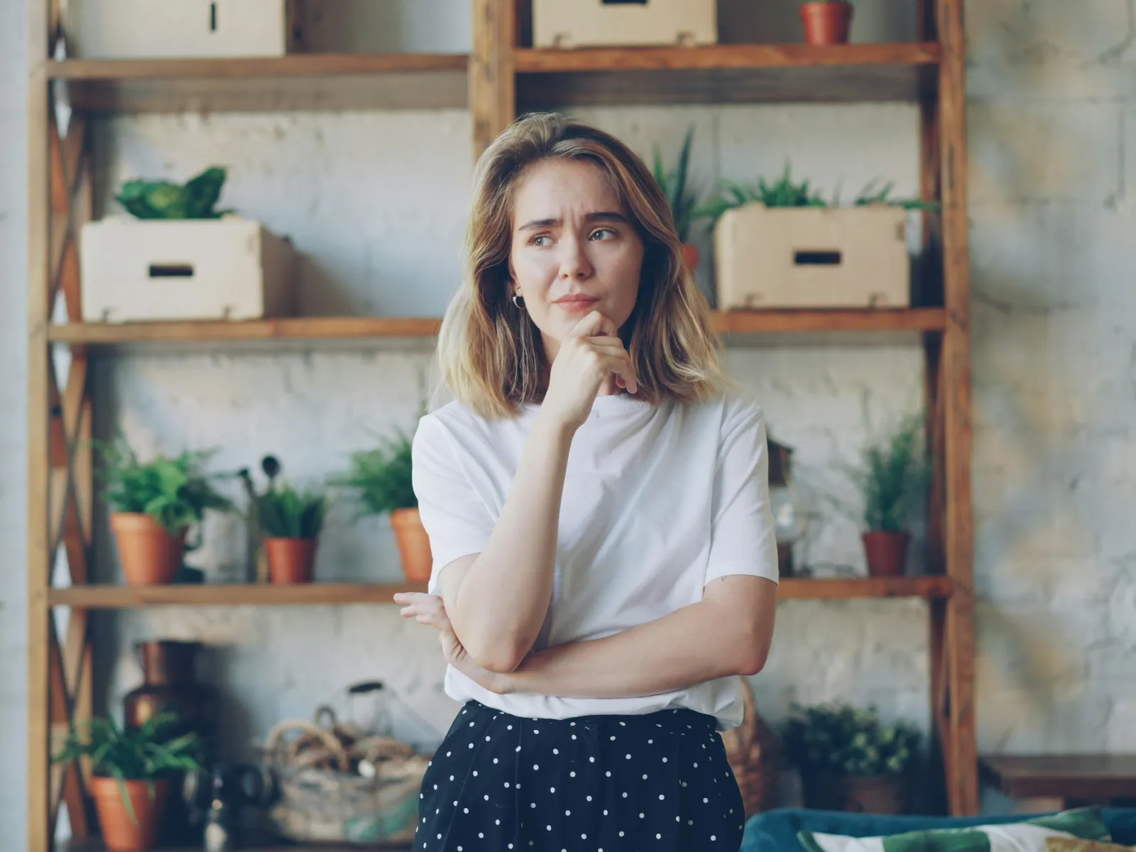 Thoughtful woman in white shirt and polka dot skirt standing indoors near wooden shelves with potted plants.