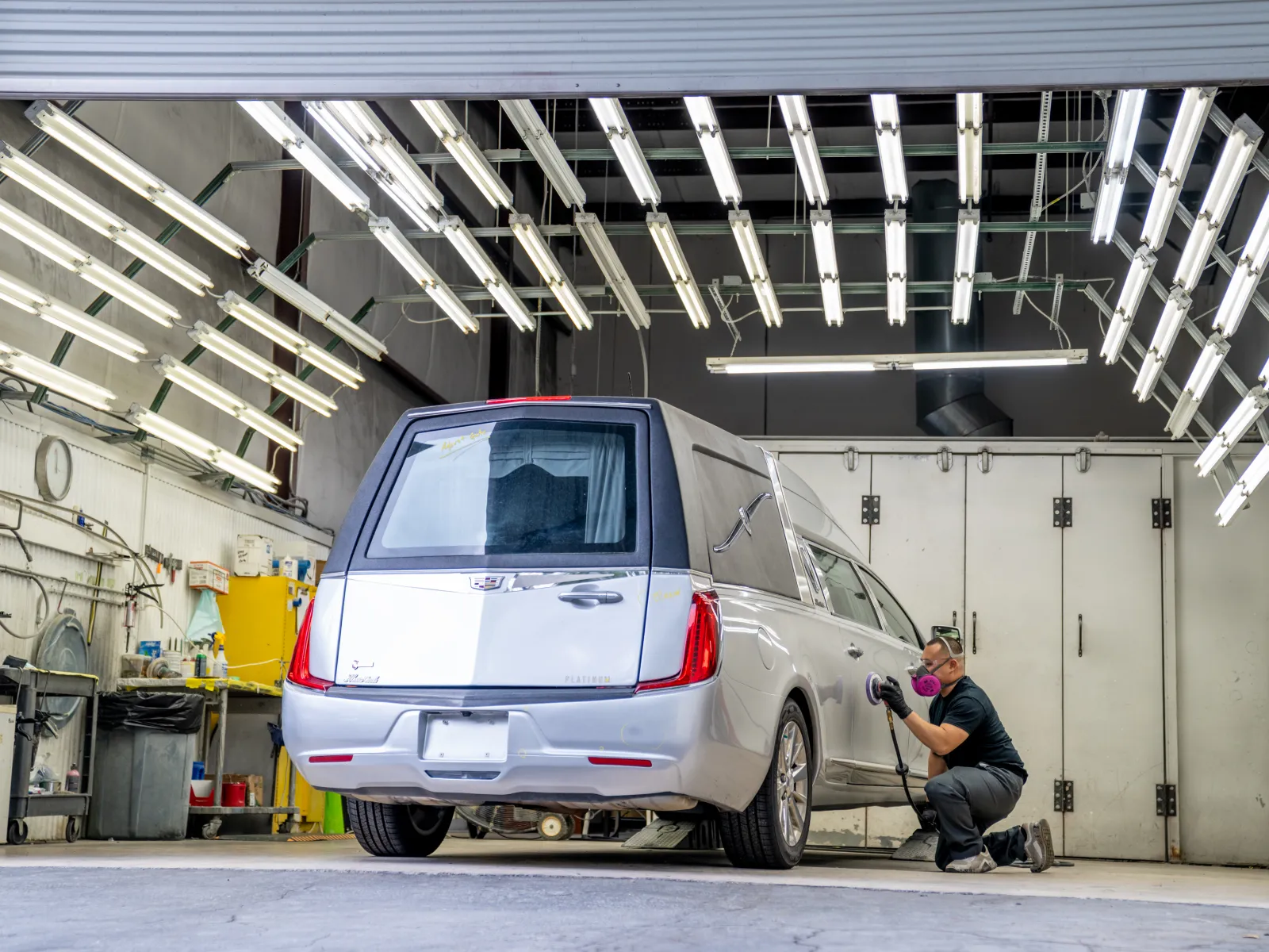 Person polishing a silver hearse inside a brightly lit workshop with numerous fluorescent lights overhead.