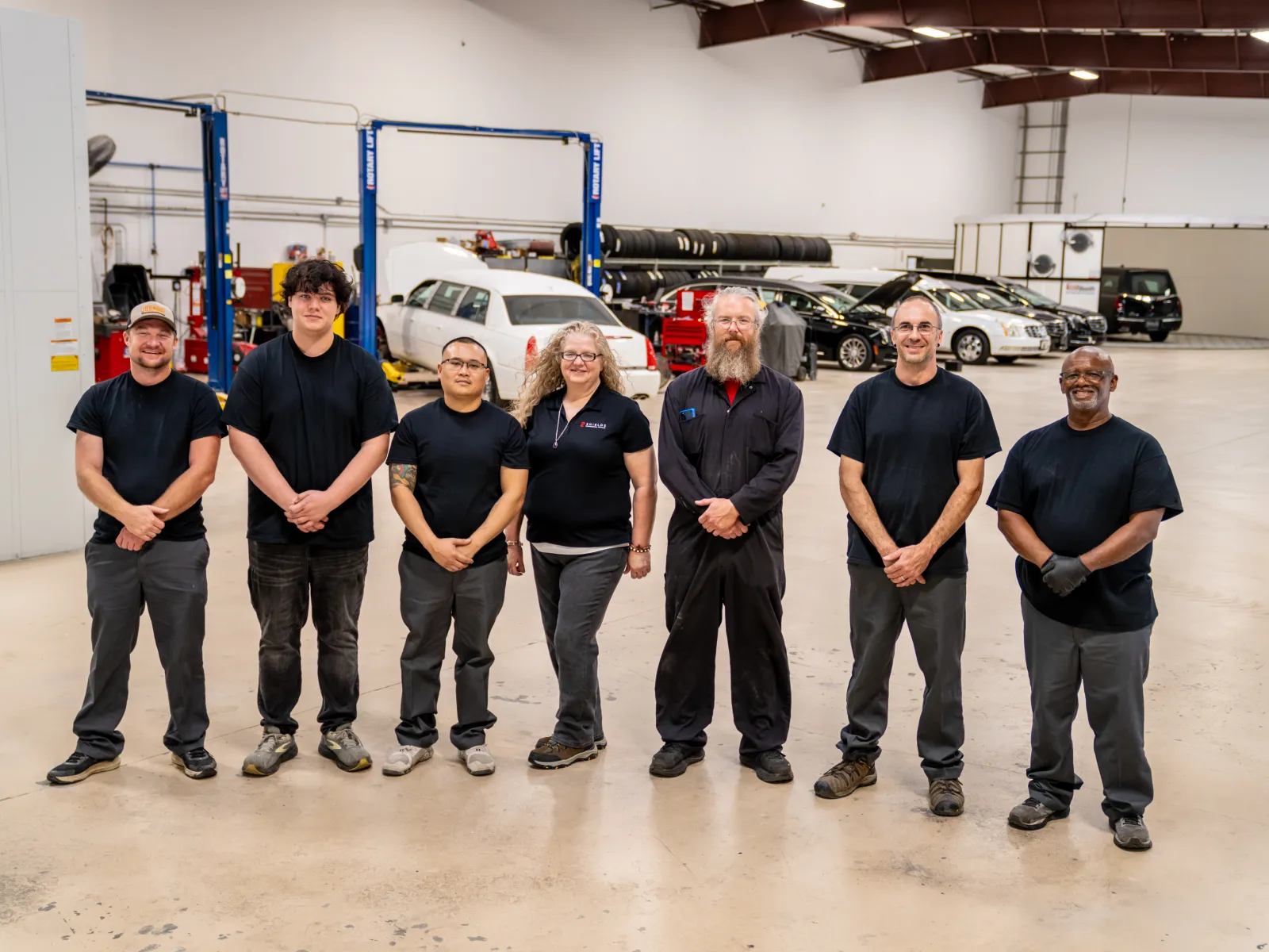 Seven auto repair technicians stand in a spacious garage with cars and equipment in the background.