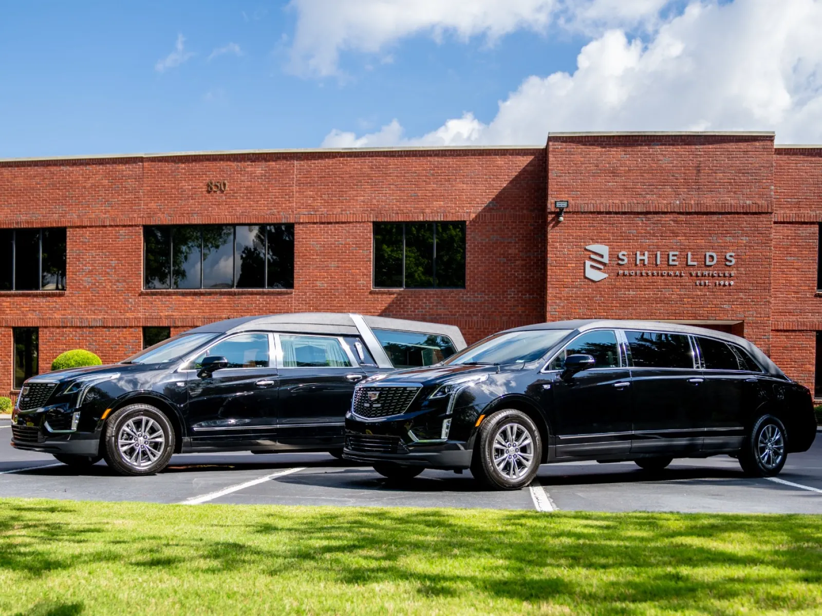 Two black luxury hearses parked in front of a brick building with a blue sky and green lawn.