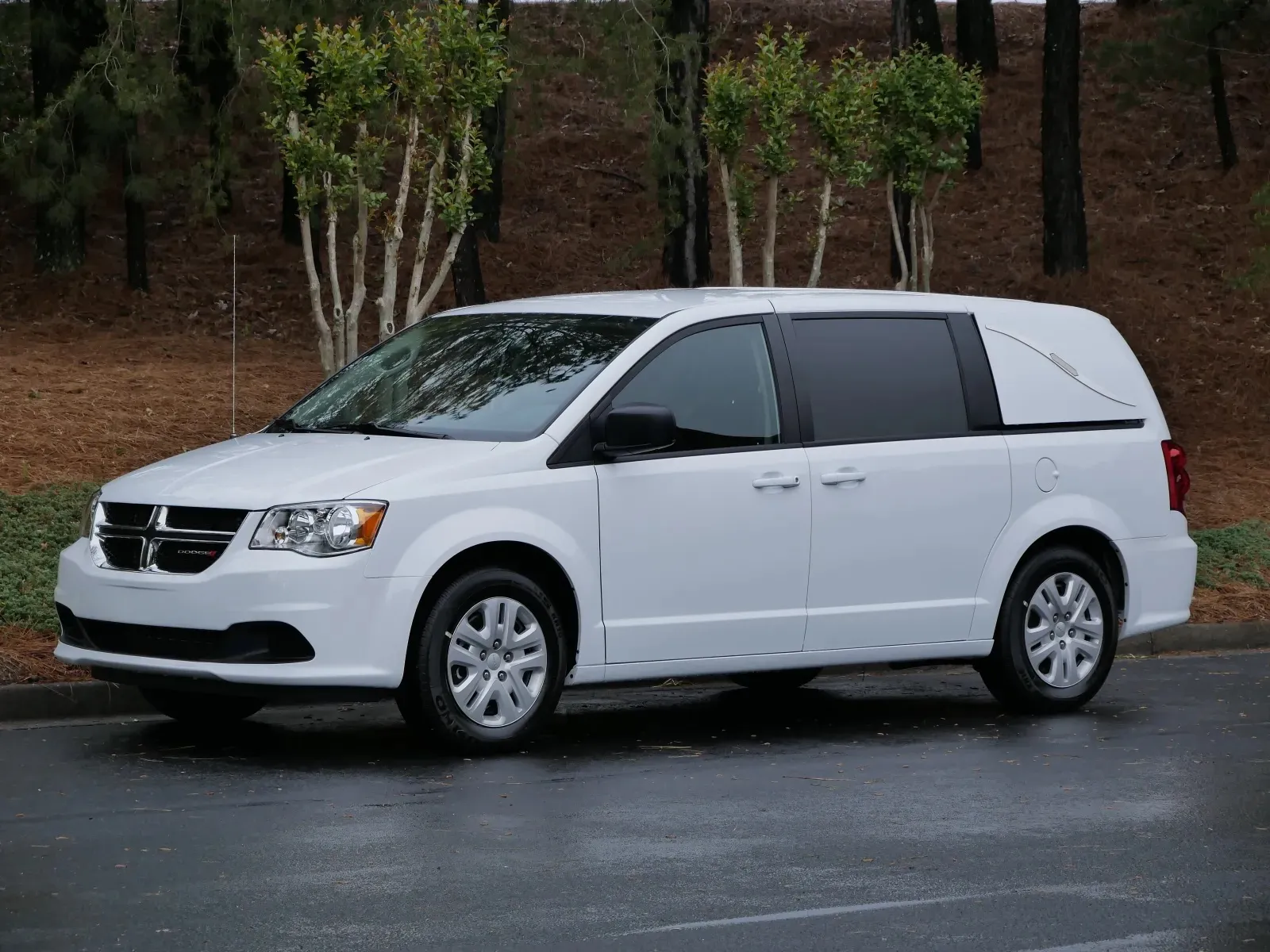 White Dodge minivan parked on a wet road surrounded by trees.