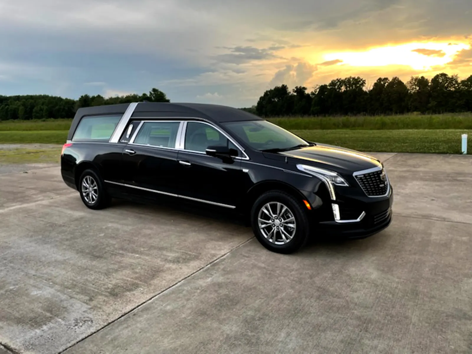 A sleek black hearse parked on a paved surface with a sunset backdrop and green fields in the distance.