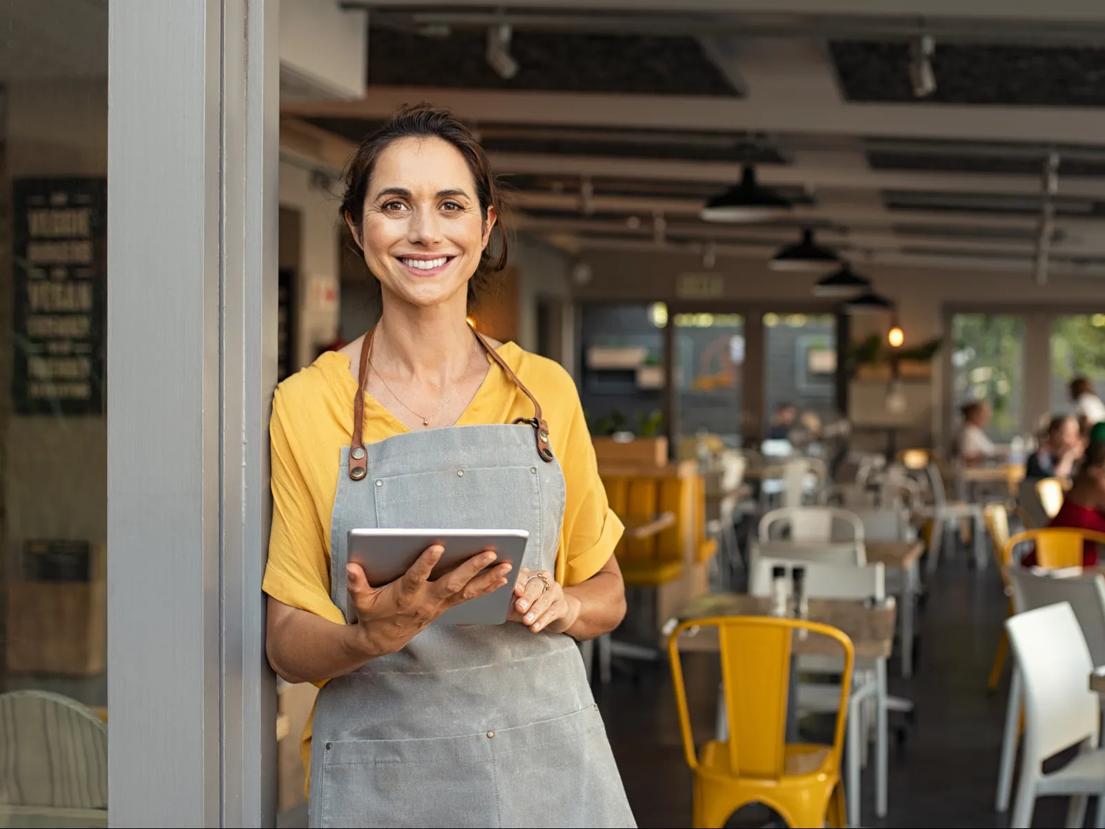 Smiling waitress in apron holding a tablet standing inside a bright restaurant with yellow chairs and customers.