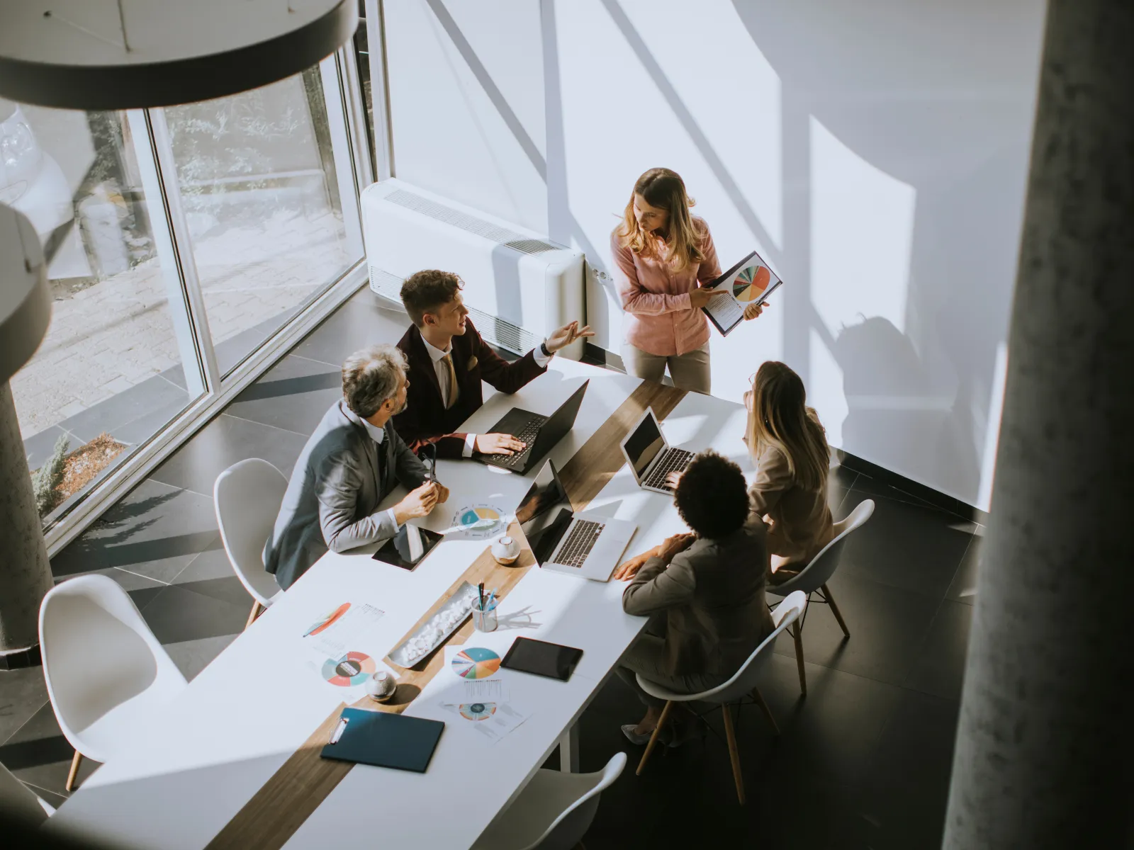 Team of five professionals collaborating in a sunlit modern office with laptops and charts on a table.