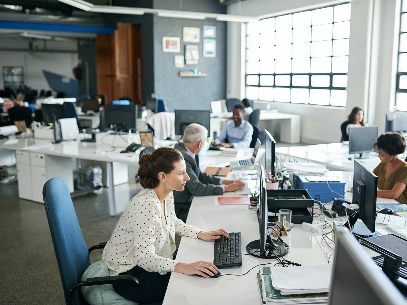 Diverse professionals working at computers in a bright, modern open-plan office with large windows.