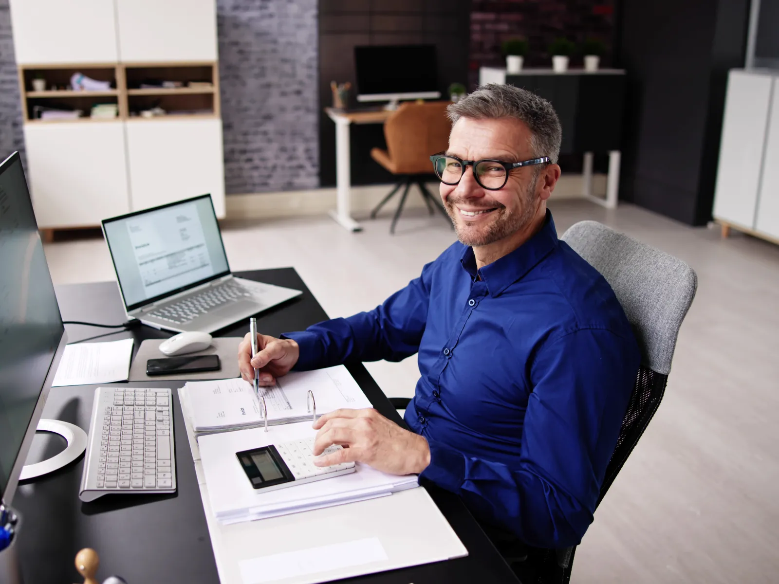 Smiling businessman in glasses using calculator and laptop while working at modern office desk