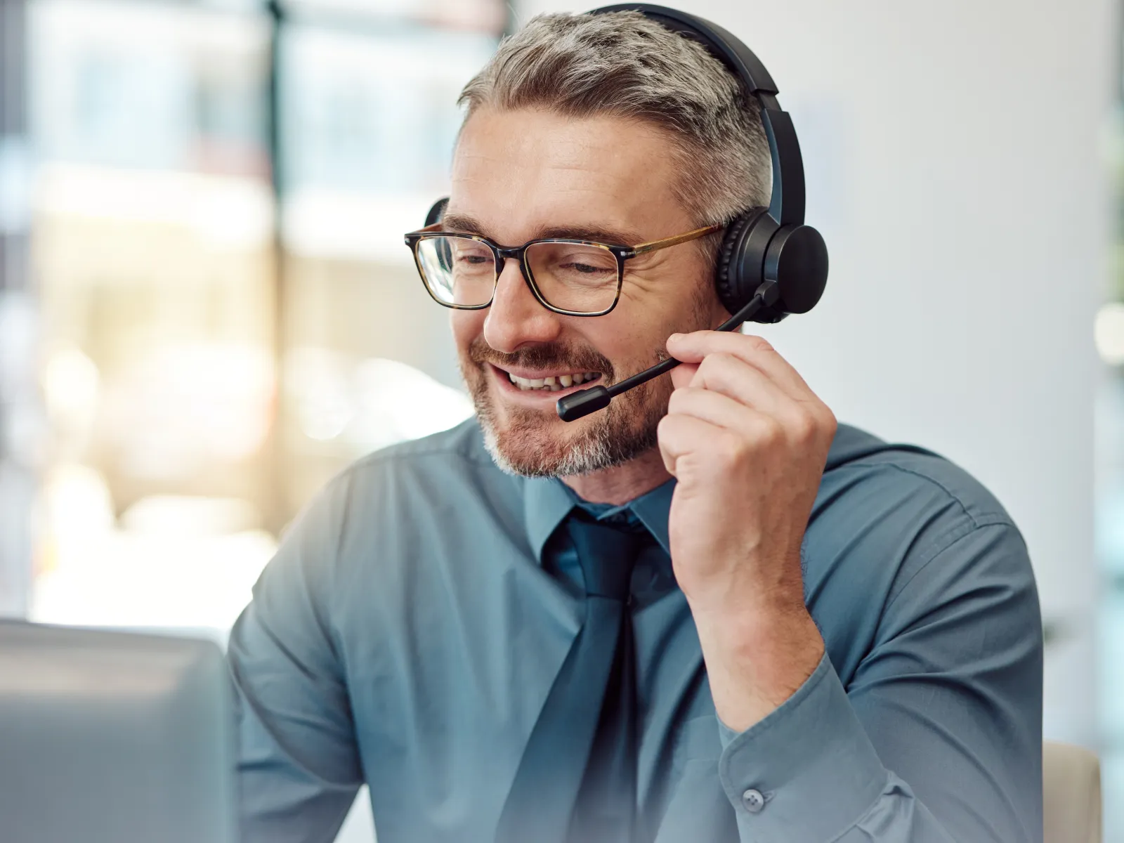 Smiling middle-aged man with headset and glasses engaged in a call at a bright office workspace