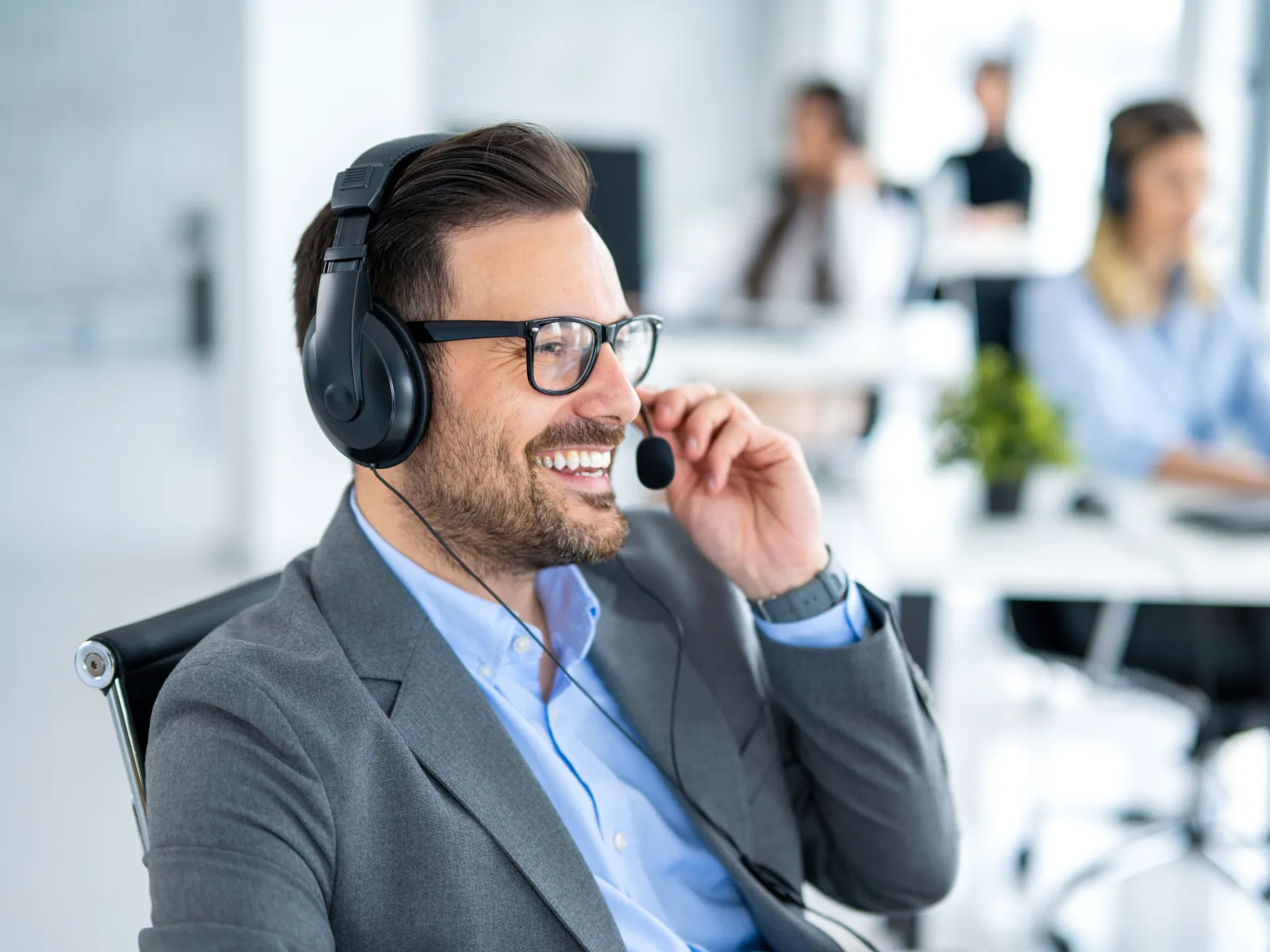 Smiling man wearing headset and glasses working in modern office with colleagues in background