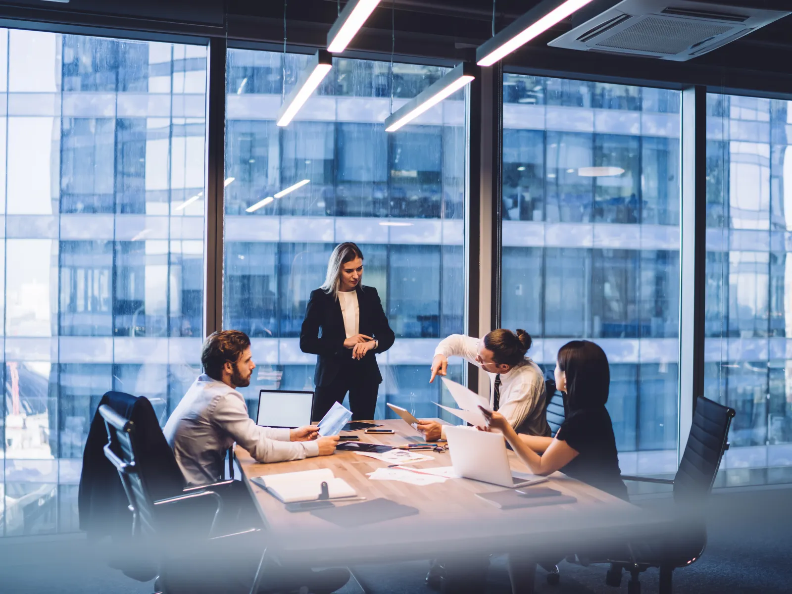 Four professionals in a modern glass office engaged in a business meeting with laptops and documents.