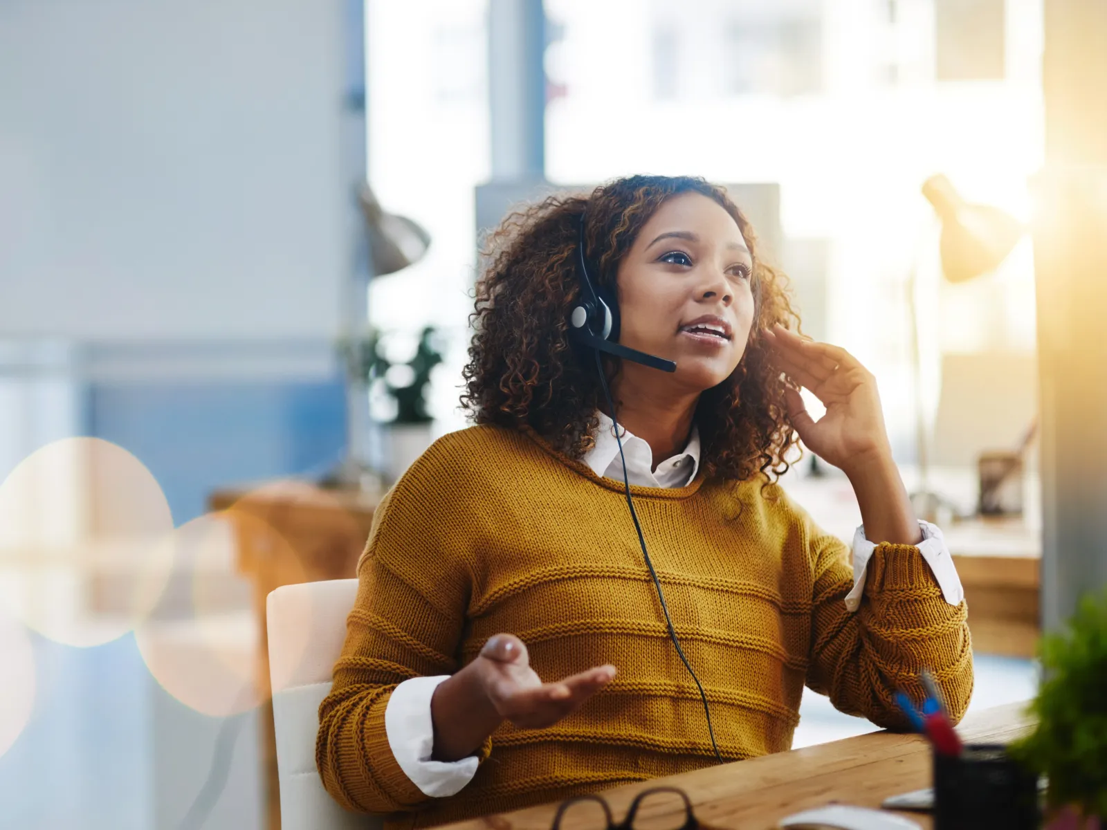 Customer service representative wearing headset and mustard sweater talking in a bright modern office.