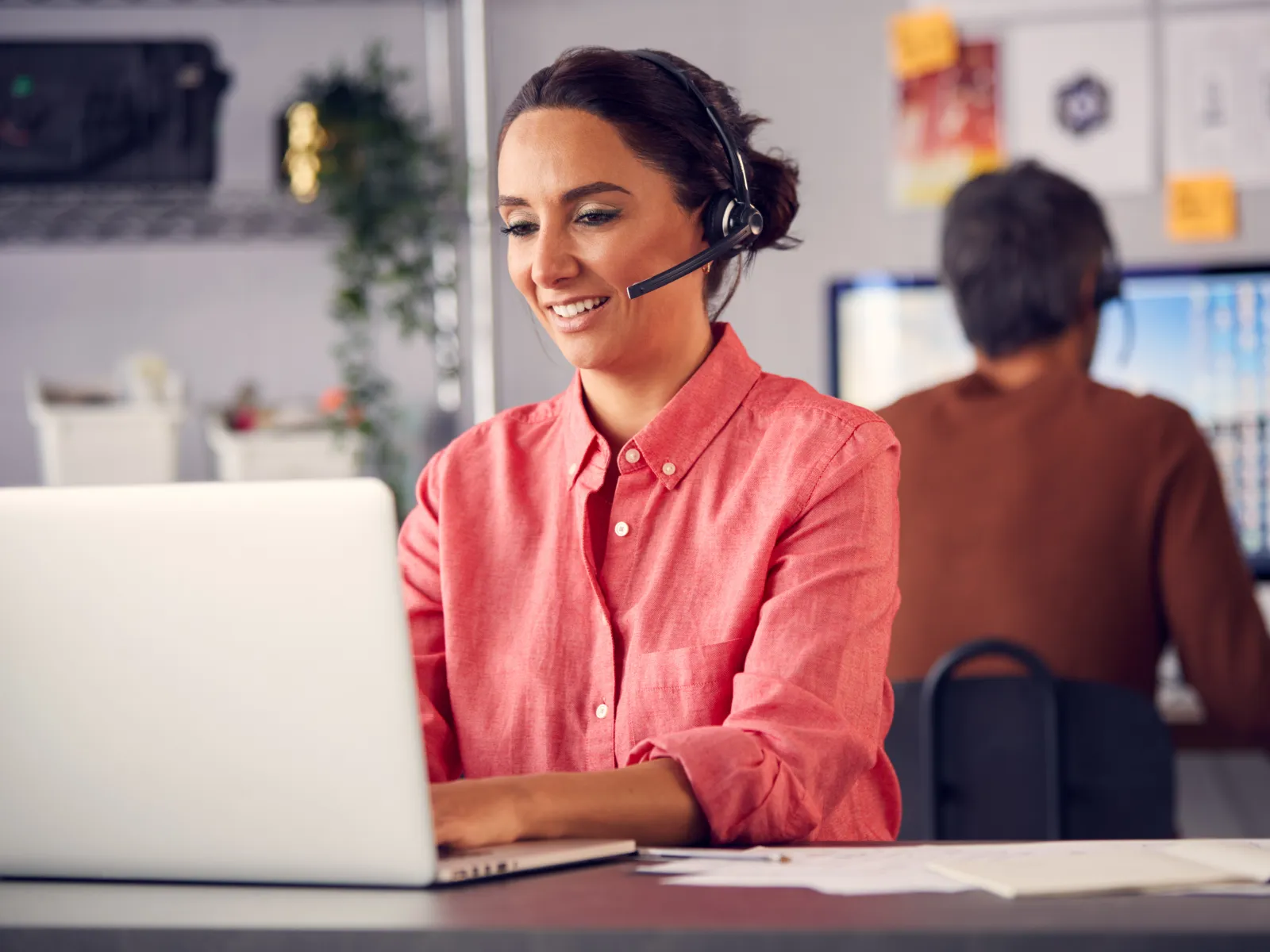 Female customer support agent wearing headset working on laptop in office with colleague at computer