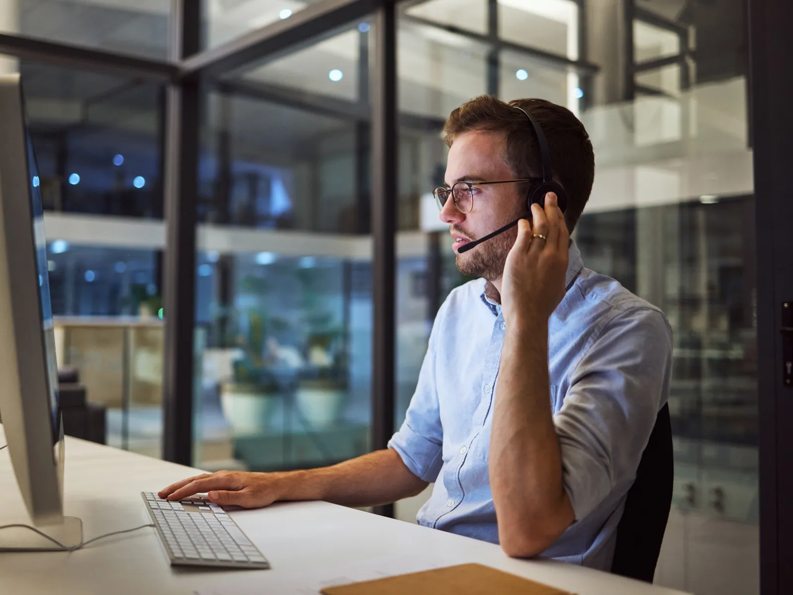 Man wearing headset working on desktop computer in modern glass office during evening hours