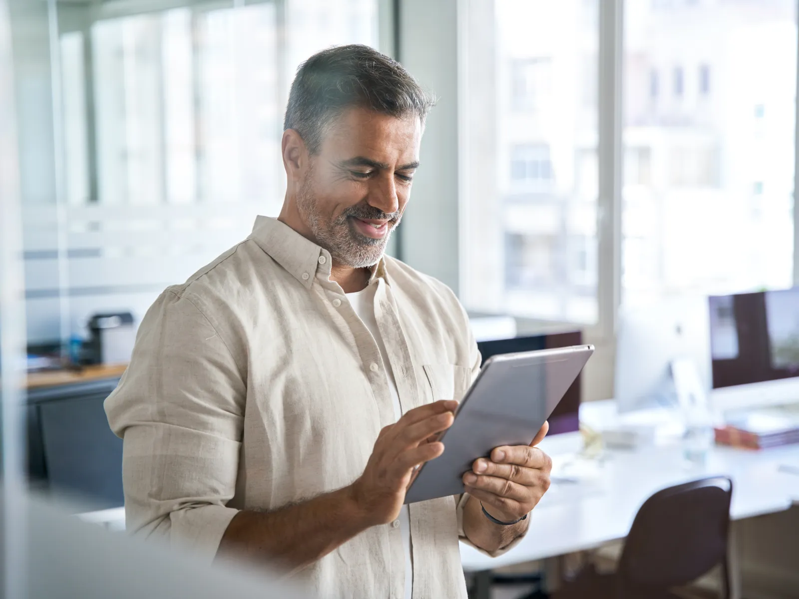 Smiling man using a digital tablet in a bright modern office space with desks and computers.