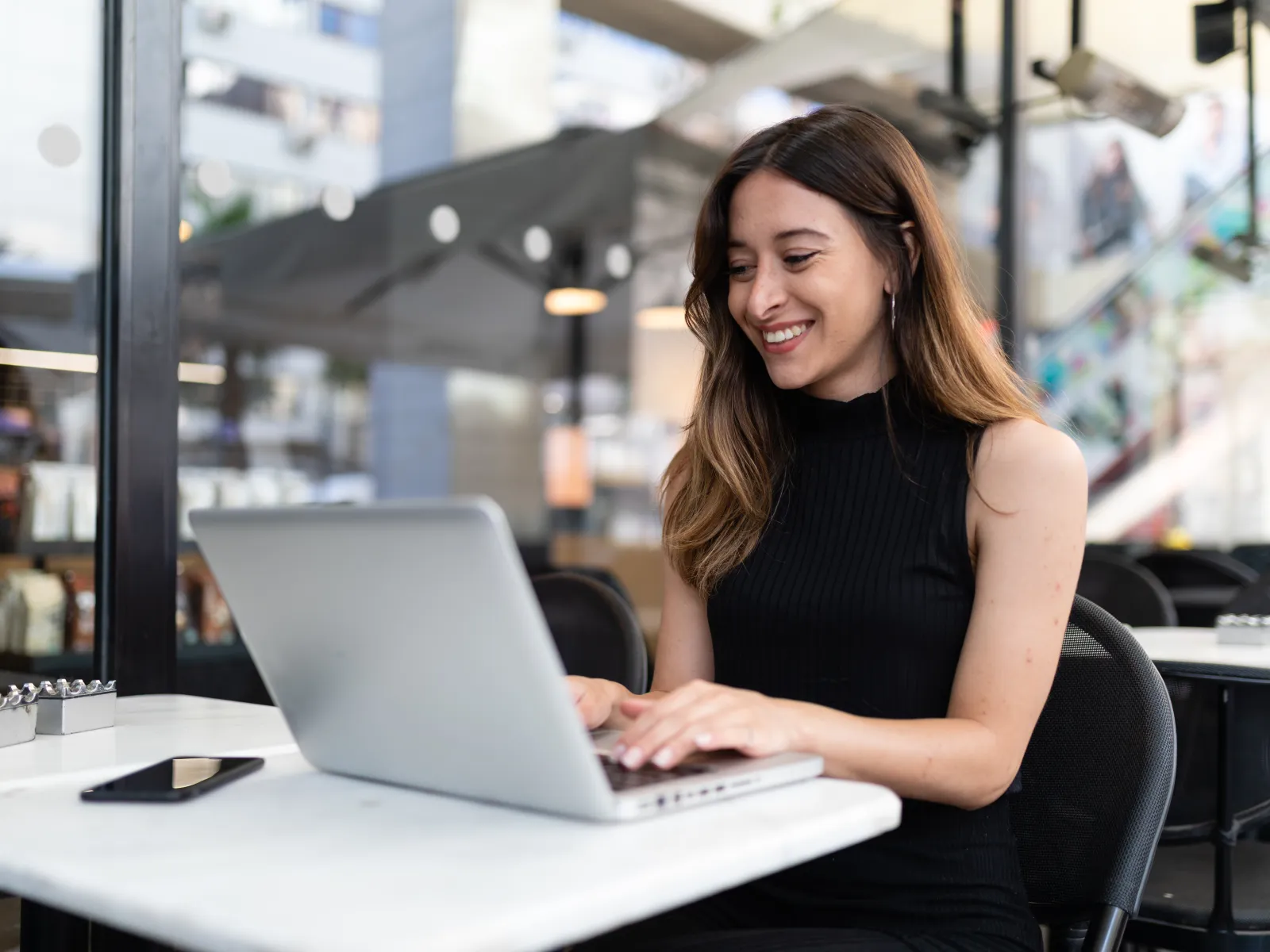Young woman smiling while working on a laptop at a cafe with a smartphone on the table